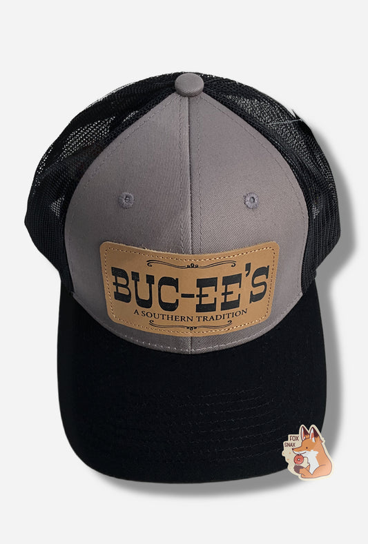 A picture of a grey and black ball cap.   The front of the cap is grey, and has a large rectangular leather patch that reads BUC-EE'S, and underneath that in much smaller text, A SOUTHERN TRADITION.  The brim of the cap is black.  The back and sides of the cap are a black plastic mesh.  A paper label is attached to the top of the cap; it reads TOP GRAIN LEATHER PATCH.