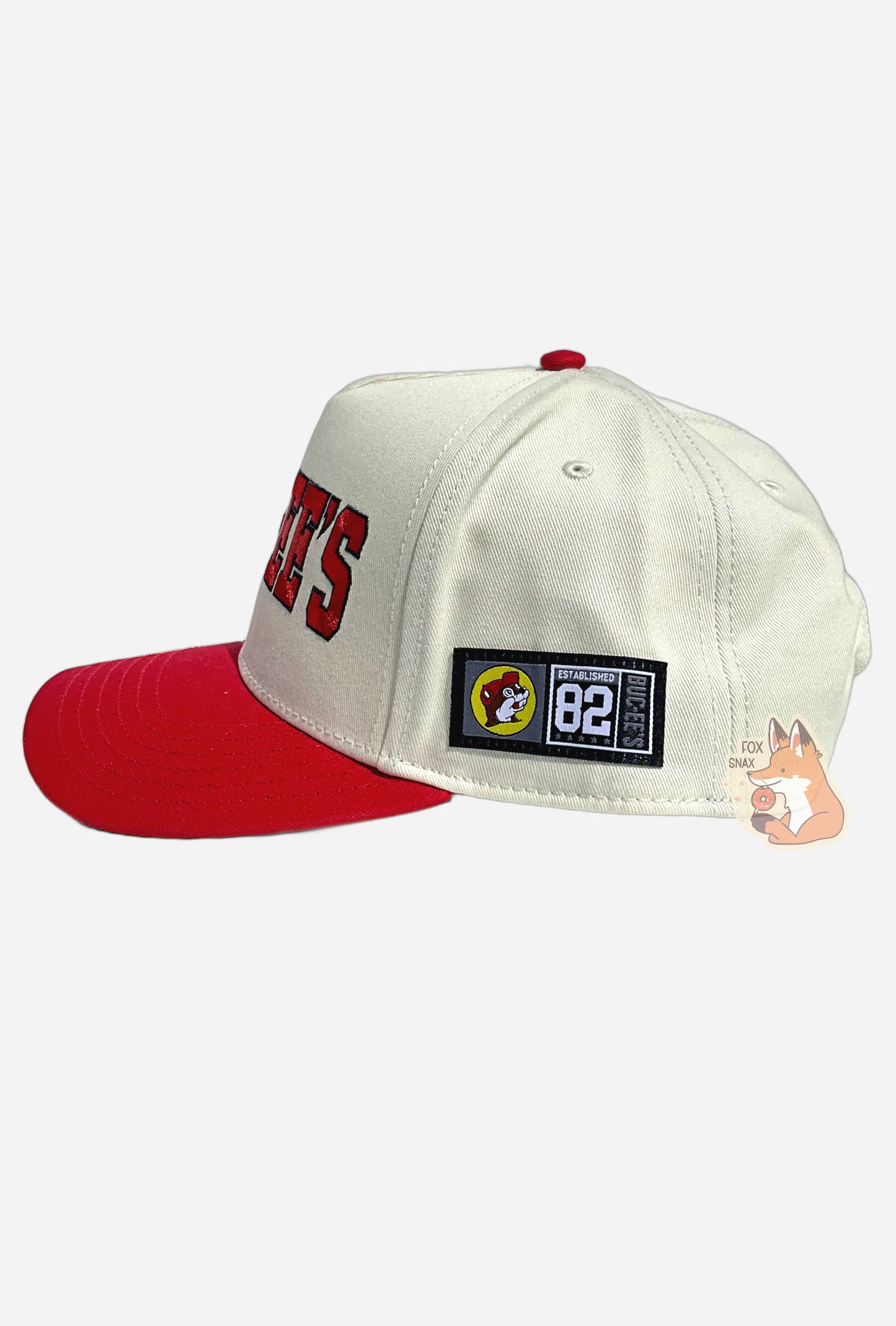 A picture of the side of a red and white baseball cap.  The body of the cap is white.  The brim of the cap is red.  The button on top of the cap and the snapback on top of the cap.  The back and sides of the cap are a white cloth.  On the side, there is a small rectangular patch that reads ESTABLISHED 82, BUC-EES, next to a Buc-ee The Beaver logo.