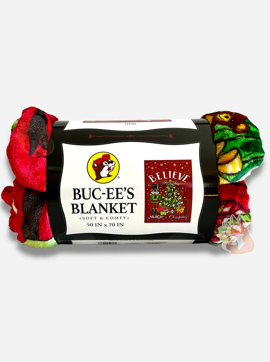 A picture of the side of a soft red, black, green, and yellow blanket featuring Buc-ee the Beaver as a nutcracker, next to a Christmas tree.  The tree has many presents underneath, including a Buc-ee's logo.  The background is in red, framed by white snowflakes at the top, and reads BELIEVE IN THE MAGIC OF CHRISTMAS.  The label reads BUC-EE'S BLANKET *SOFT AND COMFY* 50 IN x 70 IN