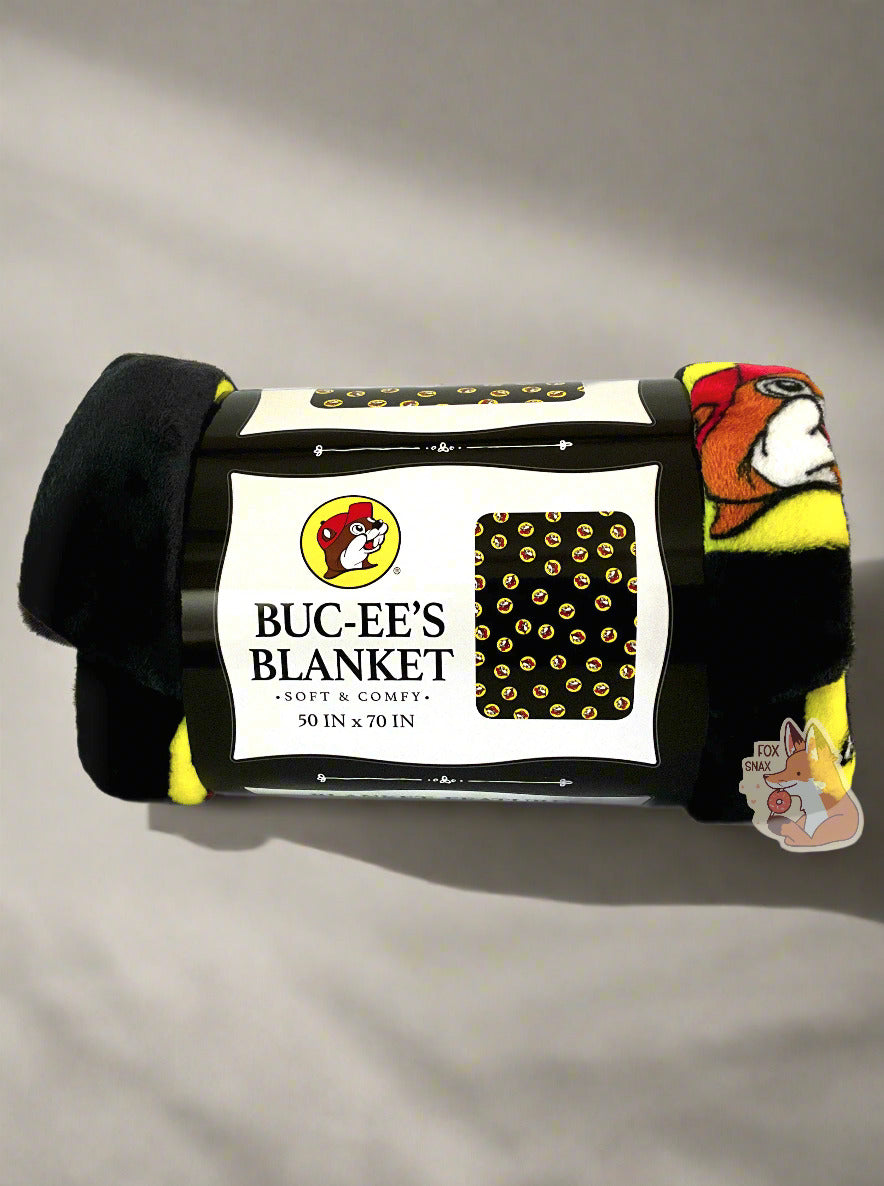 A picture of a soft black blanket covered in Buc-ee's logos.  The logo is Buc-ee The Beaver, wearing a red ball cap against a yellow circle background.  The label reads BUC-EE'S BLANKET *SOFT AND COMFY* 50 IN x 70 IN.  The background is a soft grey with shadows.
