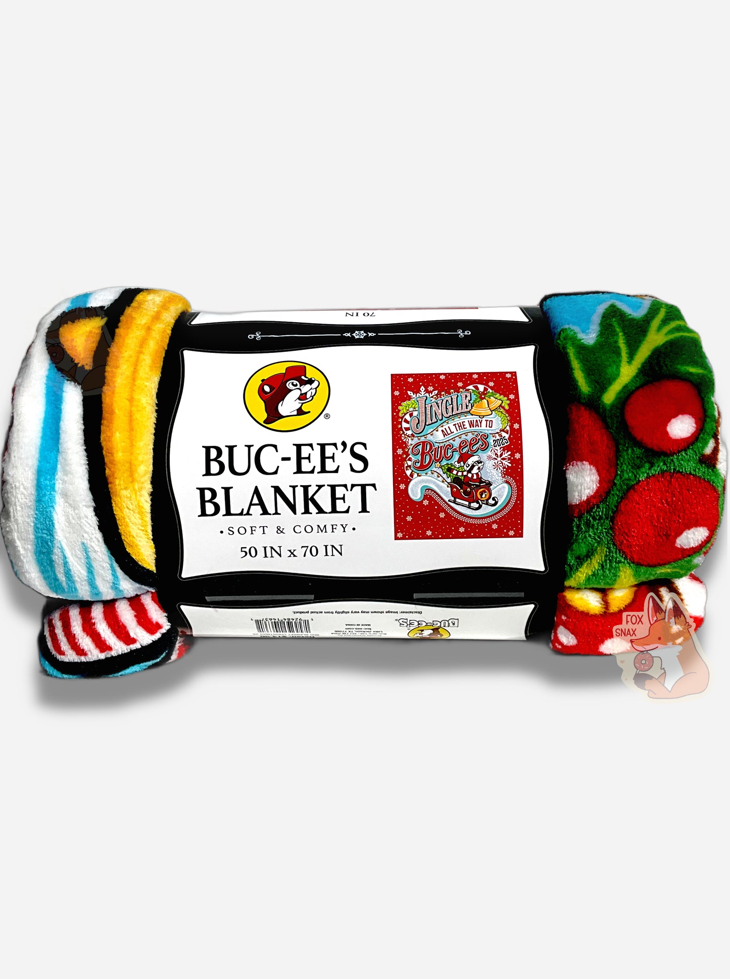 A picture of the side of a soft white, blue, blank, yellow, green, and red (!) blanket featuring Buc-ee the Beaver in a red Christmas sleigh, with a green Christmas tree in the back.  Buc-ee is holding the front of the sleigh, and is wearing a Christmas cap and red and white striped scarf.  The background is in red, framed by white snowflakes at the top, and reads JINGLE ALL THE WAY TO BUC-EE'S.  The label reads BUC-EE'S BLANKET *SOFT AND COMFY* 50 IN x 70 IN
