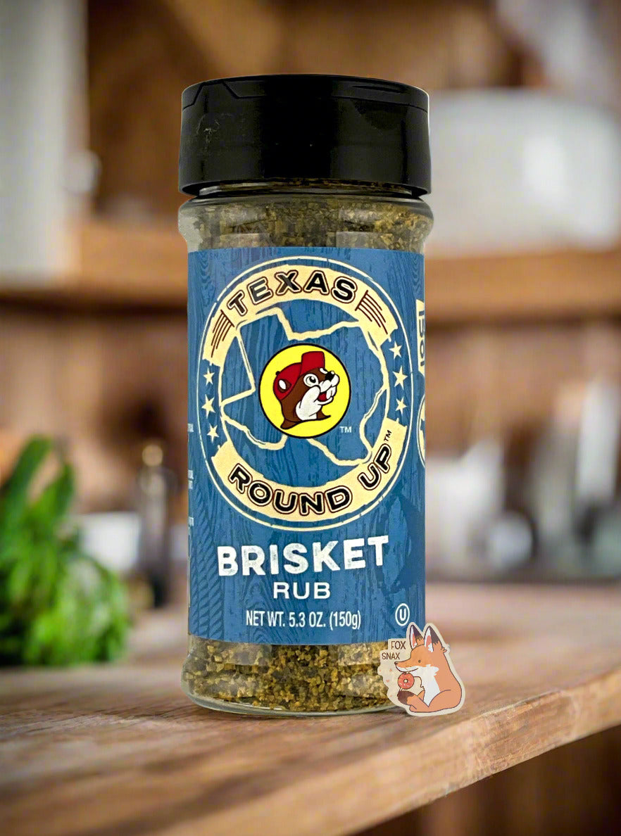 A picture of the front of a container of Texas Round Up Brisket Rub.  The container has a black plastic flip lid, and a clear plastic body.  The spices inside appear in shades of grey and black.  The label reads TEXAS ROUND UP, with an outline of the state of Texas, with a picture of Buc-ee The Beaver in the middle.  Beneath that, the blue label reads in white text, Brisket Rub.  Net Wt 5.3 Oz (150g).  The background shows an amazing, rustic, wood-paneled kitchen.