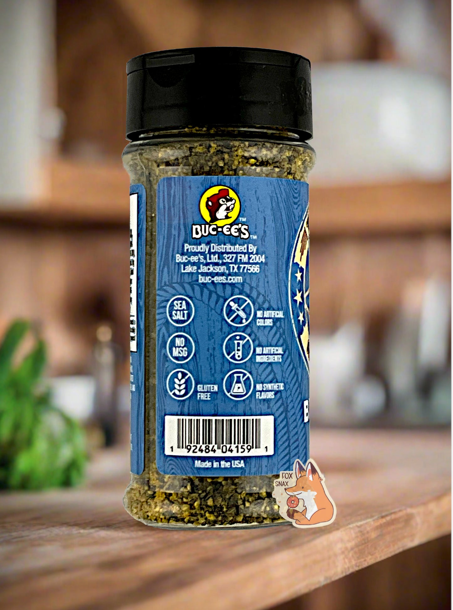 A picture of the side of a container of Texas Round Up Brisket Rub.  The container has a black plastic flip lid, and a clear plastic body.  The spices inside appear grey and black.  The blue label reads SEA SALT, NO MSG, GLUTEN FREE, NO ARTIFICIAL COLORS, NO ARTIFICIAL INGREDIENTS, NO SYNTHETIC FLAVORS.  Beneath that, MADE IN THE USA.  The background shows an amazing, rustic, wood-paneled kitchen.