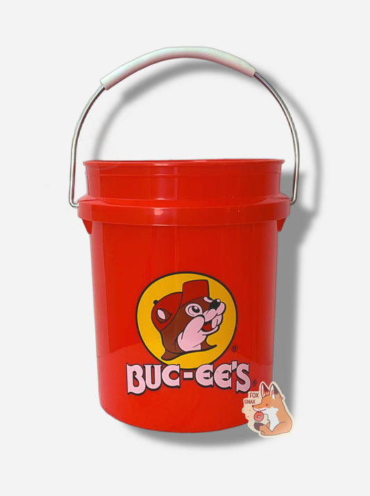 A small, bright red, miniature bucket.  The bucket has the Buc-ee's logo on it - a cheerful beaver wearing a red hat, framed by a yellow circle.  The words BUC-EE'S are underneath the logo in white.  A metal handle, partially coated in white plastic, is attached to the top of the bucket.