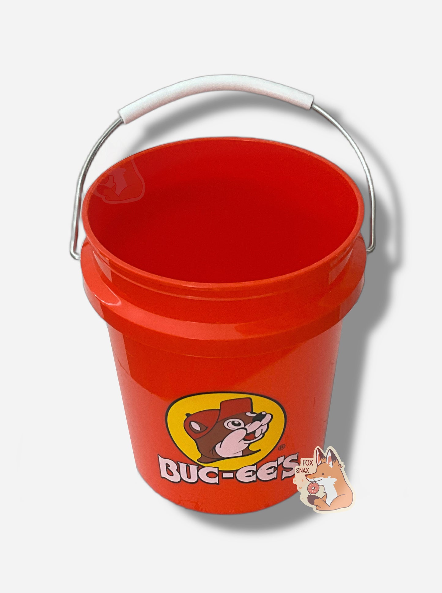 A small, bright red, miniature bucket in three quarters view.  The bucket has the Buc-ee's logo on it - a cheerful beaver wearing a red hat, framed by a yellow circle.  The words BUC-EE'S are underneath the logo in white.  A metal handle, partially coated in white plastic, is attached to the top of the bucket.