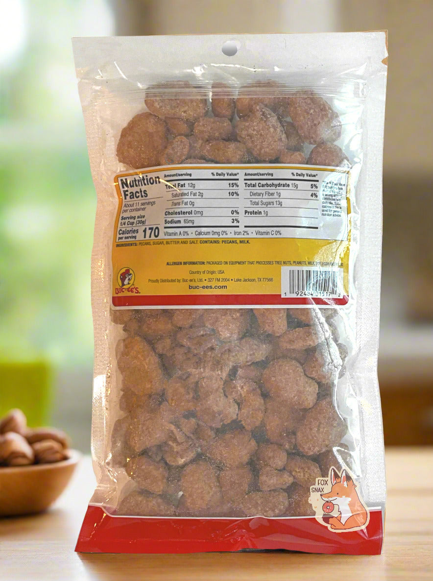 A picture of the back of a clear plastic bag of Buc-ee's Butter Toffee Pecans.  Inside the bag are a large number of dried pecans, covered in brownish butter toffee  material.  The bag is sitting on a light wood countertop, framed by a kitchen in the background; a bowl of nuts can be seen the background.