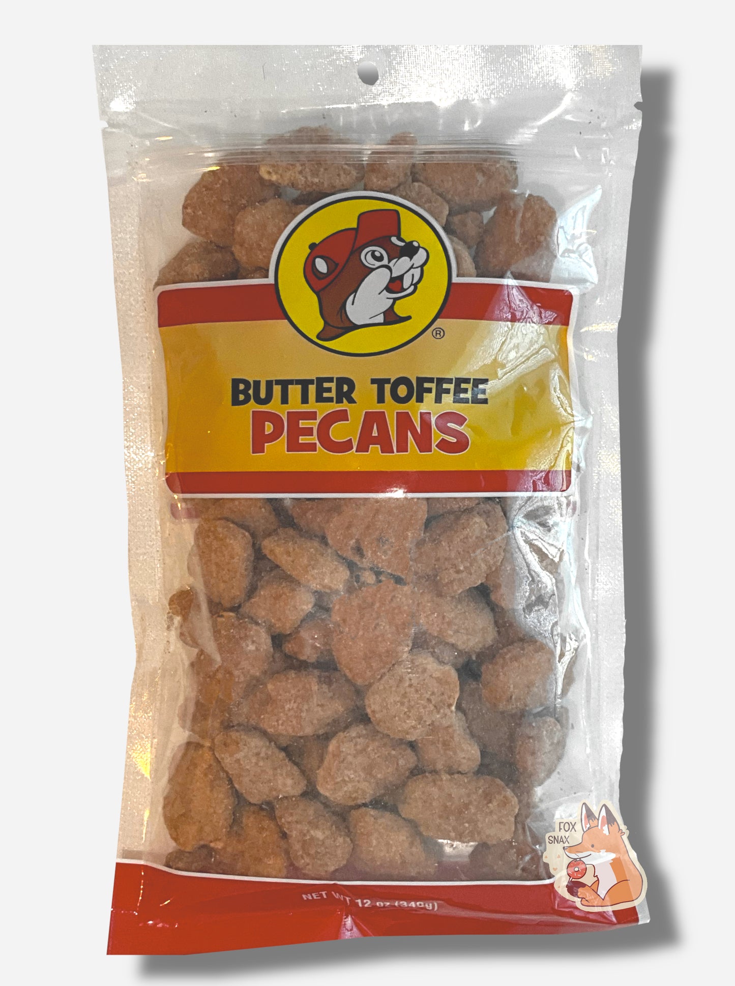 A picture of a clear plastic bag of Buc-ee's Butter Toffee Pecans.  The Buc-ee's logo can be seen in the top center of the bag, then underneath that in black and red text, BUTTER TOFFEE PECANS.  The bottom of the bag is red and reads Net Wt 12 oz.  Inside the bag are a large number of brown butter toffee pecans, covered in brownish sugary material.