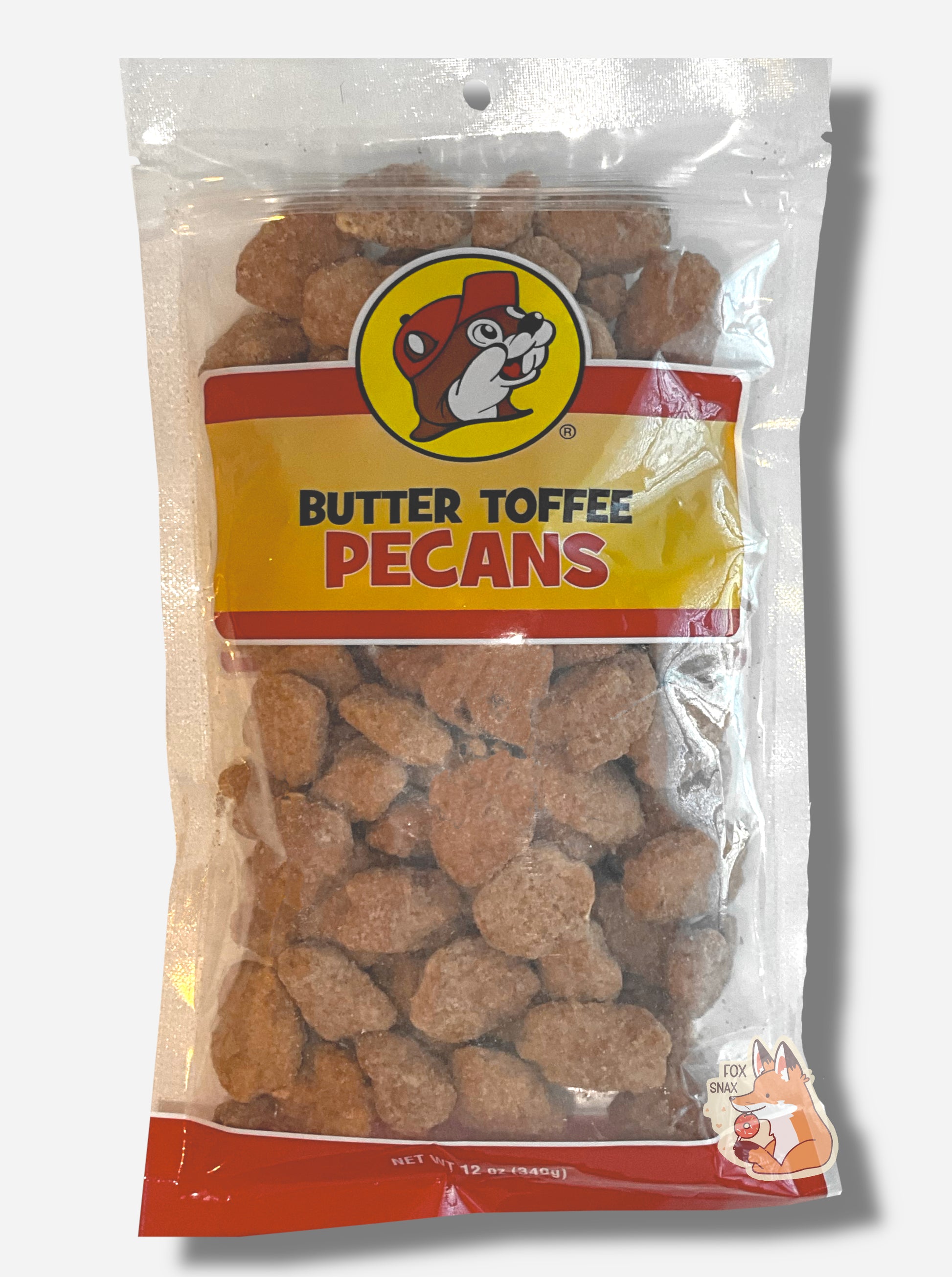 A picture of a clear plastic bag of Buc-ee's Butter Toffee Pecans.  The Buc-ee's logo can be seen in the top center of the bag, then underneath that in black and red text, BUTTER TOFFEE PECANS.  The bottom of the bag is red and reads Net Wt 12 oz.  Inside the bag are a large number of brown butter toffee pecans, covered in brownish sugary material.