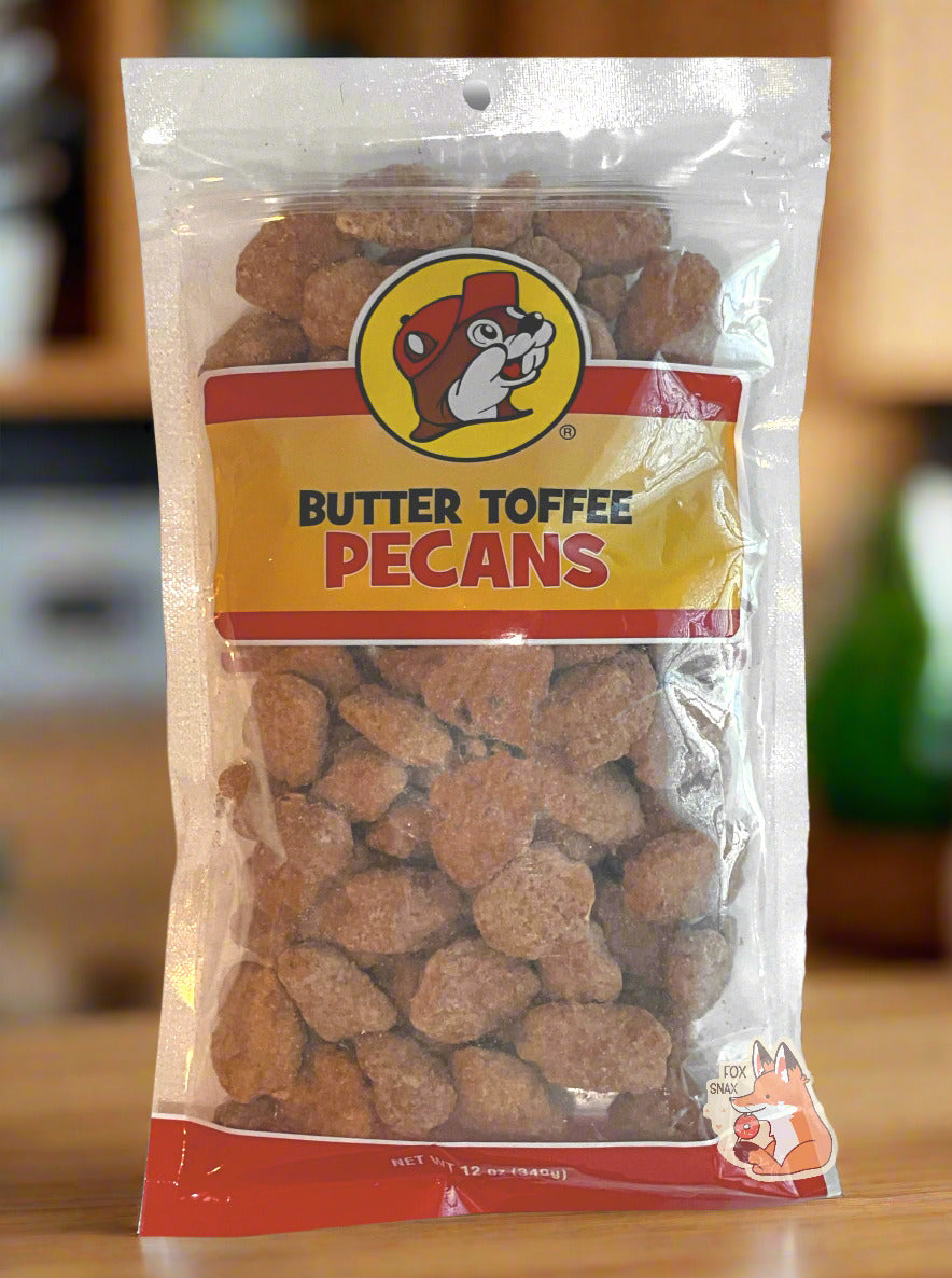 A picture of a clear plastic bag of Buc-ee's Butter Toffee Pecans.  The Buc-ee's logo can be seen in the top center of the bag, then underneath that in black and red text, BUTTER TOFFEE PECANS.  The bottom of the bag is red and reads Net Wt 12 oz.  Inside the bag are a large number of brown butter toffee pecans, covered in brownish sugary material.  The bag is sitting on a light wood countertop, framed by a kitchen in the background.