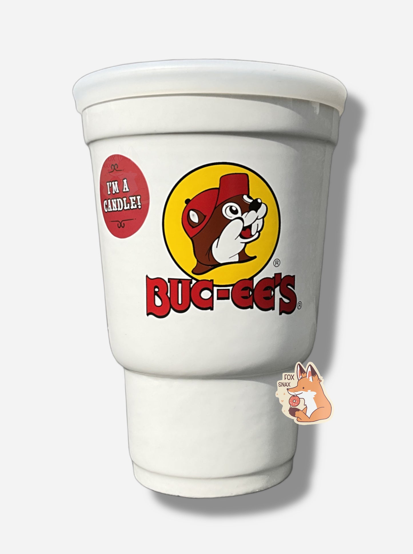 A picture of the front  of a large white ceramic candle holder, made to look exactly like a Buc-ee's stryofoam drink cup.  The surface of the cup has the Buc-ee's logo: a cheerful brown-furred beaver with a massive overbite, wearing a ball cap, and underneath it in red text, BUC-EE'S.  A small removable circular red sticker on the side of the cup reads i'M A CANDLE!