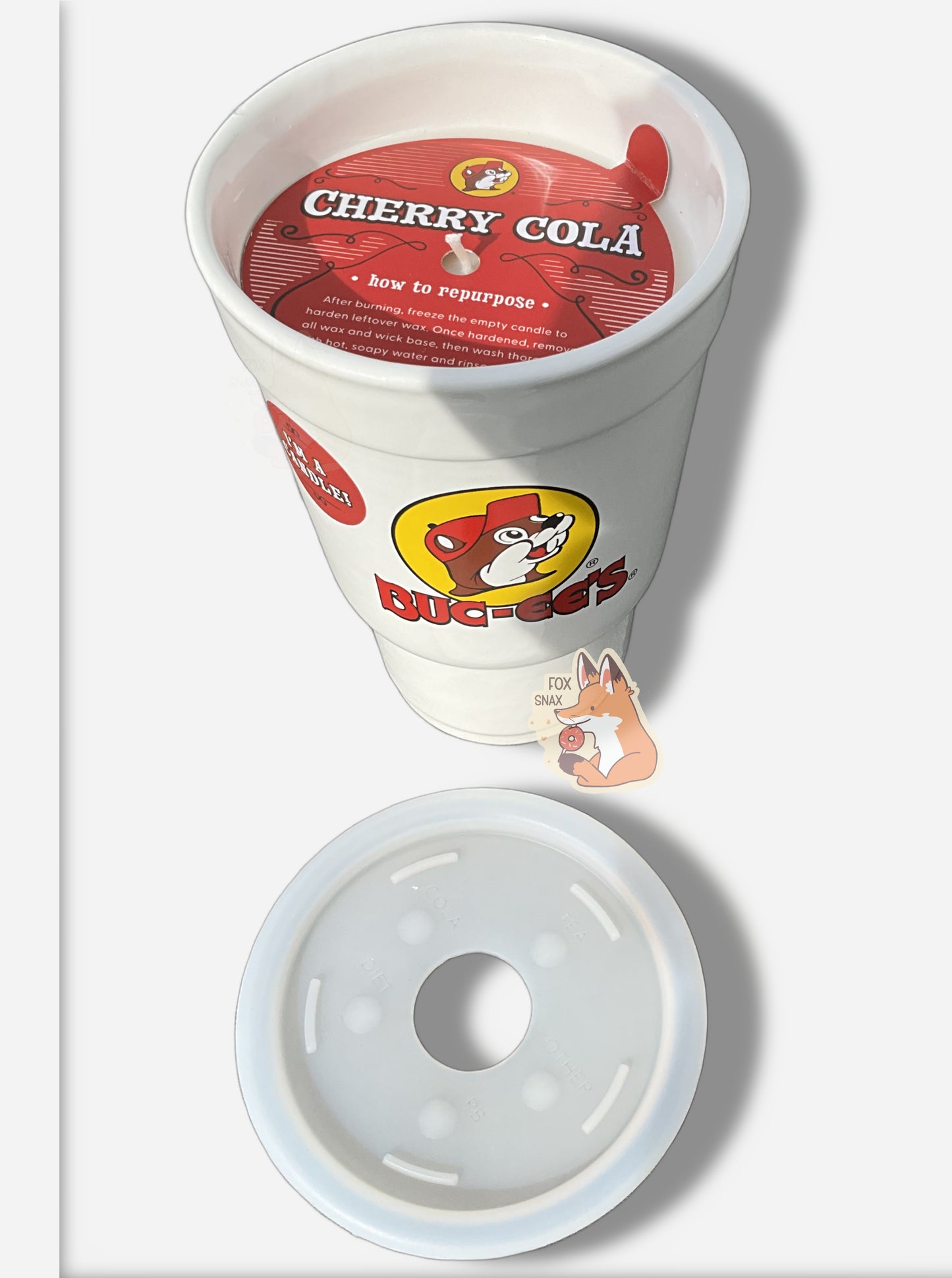 A three-quarters view of the top and front  a large white ceramic candle holder, made to look exactly like a Buc-ee's stryofoam drink cup.  The surface of the cup has the Buc-ee's logo: a cheerful brown-furred beaver with a massive overbite, wearing a ball cap, and underneath it in red text, BUC-EE'S.  The top of the candle, removed an put nearby, is a flexible rubbery material..  Inside the candle you can see white wax, a wick, and the words CHERRY COLA followed by reusability instructions.