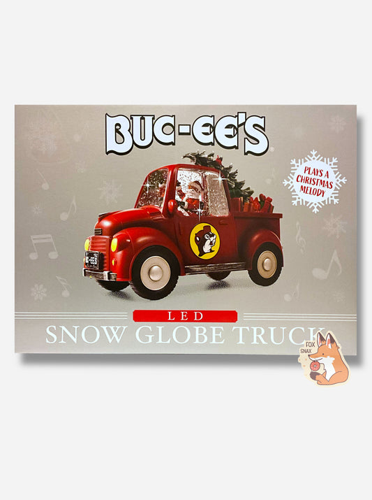 A picture of a grey rectangular box containing Buc-ee's Led Snow Globe Truck.  The front has a picture of a cartoon-ish looking red pickup truck with a black Buc-ee's license plate.  The inside of the truck has Buc-ee The Beaver, wearing a Santa hat, with his right arm upraised in a wave.  Surrounding Buc-ee is glitter (it's a snow globe).  The back of the truck has a Christmas tree and presents galore.  A snowflake in the background reads PLAYS A CHRISTMAS MELODY.