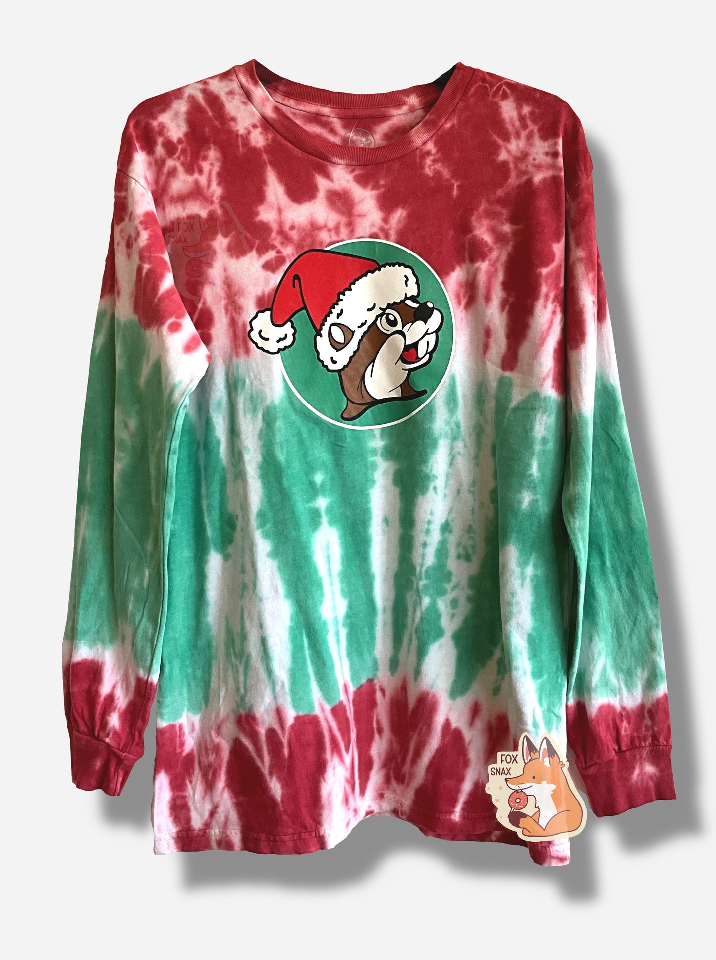 A picture of a red, white, and green Christmas tie-dye long sleeve shirt.  The top of the shirt - sleeves, upper chest - is mostly red, with some white streaks.  A small band of uneven white is around the upper/middle chest, which fades down into green at the bottom around the belly.  In the middle is the Buc-ee's logo, but the background is green, and Buc-ee the Beaver is wearing a red Santa hat with a white pom on the end.  The bottom of the shirt shows another brief band of white, then red.