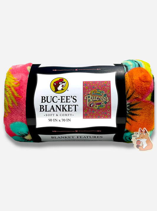 A picture of a rolled up blanket.  The blanket is a multicolored (pink, yellow, orange, black, green/blue) blanket with pastel florals and a wave of soothing colors through the center. The iconic beaver makes an appearance, rocking a pink cap, with curvy white Buc-ee's text to match.  The paper label surrounding the blanket reads BUC-EE'S BLANKET *SOFT AND COMFY* 50 IN x 70 IN