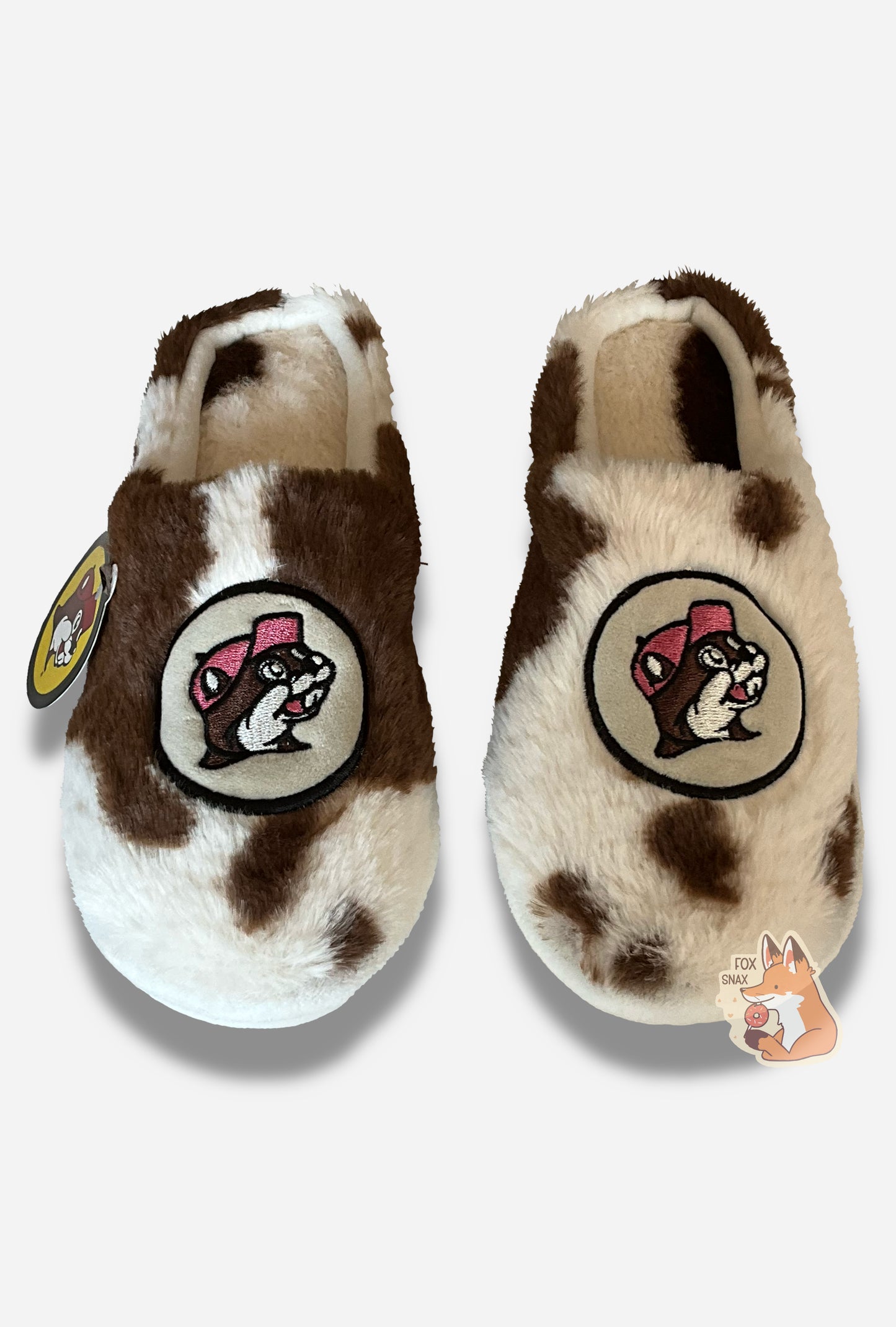 A front angled view of two cozy, fuzzy slippers.  The slippers are covered in a brown and white "fur" spotted cow print.  On the front top of each slipper, where the toe is, is the Buc-ee's logo with a cheerful beaver grin.