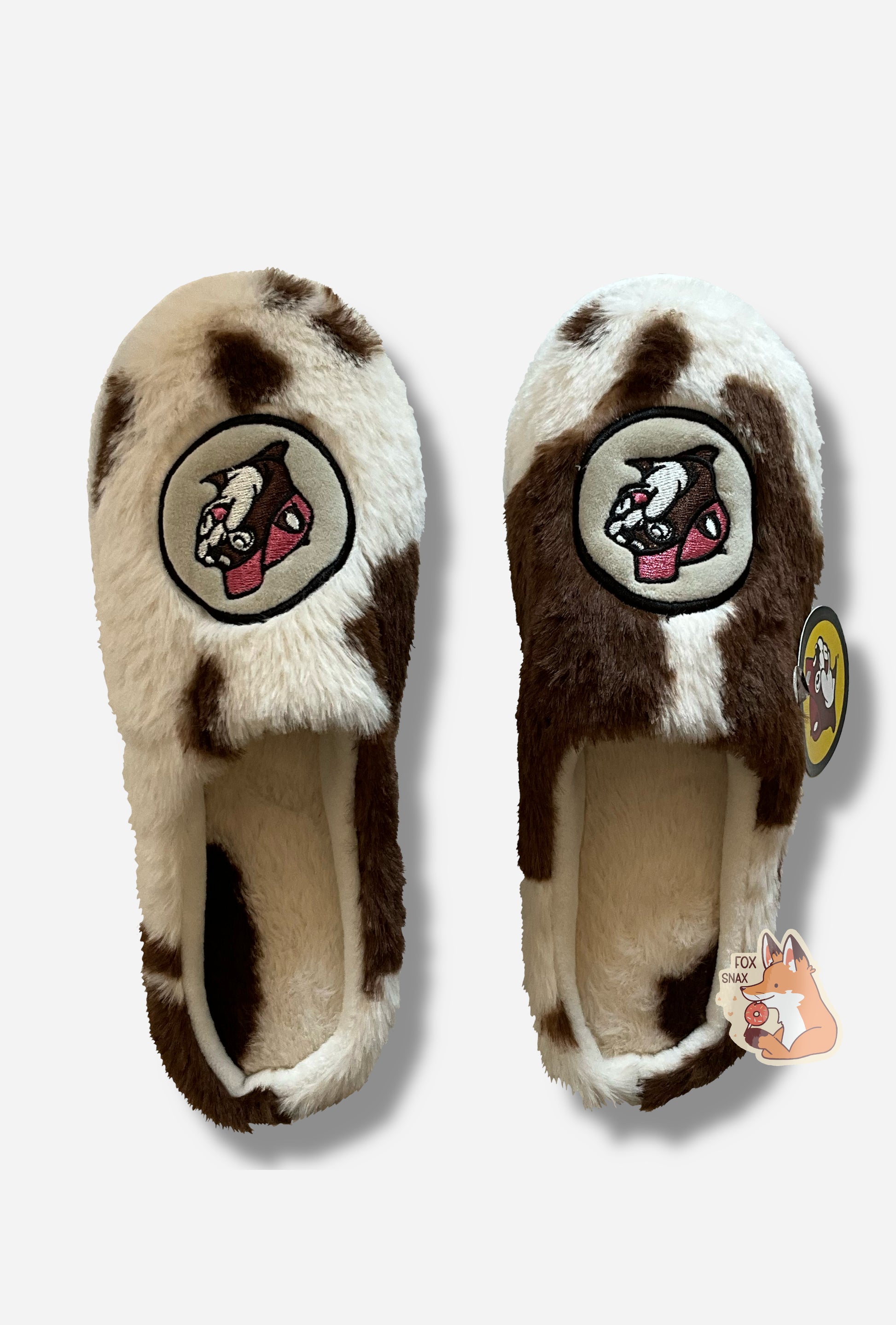 A front angled view of two cozy, fuzzy slippers.  The slippers are covered in a brown and white "fur" spotted cow print.  On the front top of each slipper, where the toe is, is the Buc-ee's logo with a cheerful beaver grin.  Looking into the top of the slipper, one can see that the cow spotted pattern extends to the inside.