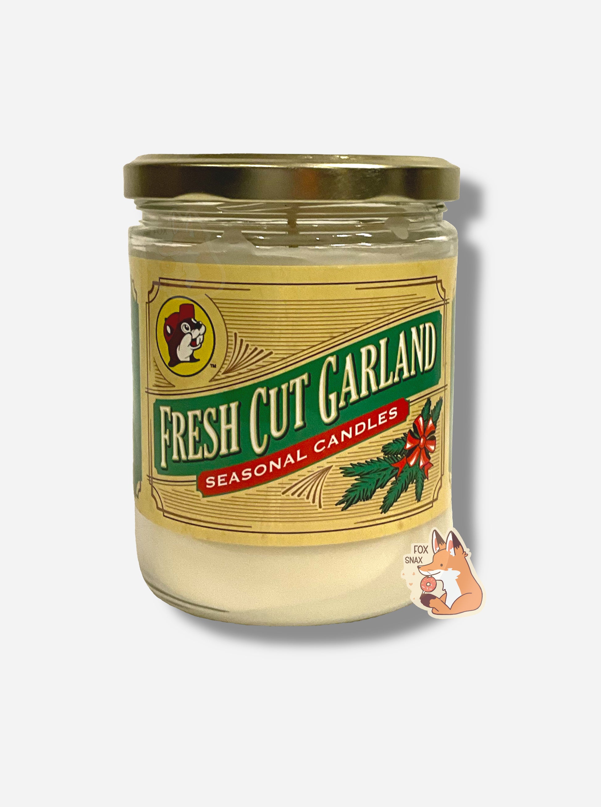 A picture of a white/yellow candle in a cylindrical glass container.  The lid of the container is a gold colored metal lid.  The front of the candle has a yellow sticker with the Buc-ee's logo, then underneath that, FRESH CUT GARLAND, then SEASONAL CANDLES, then a picture of a small fir garland with a red bow on it.