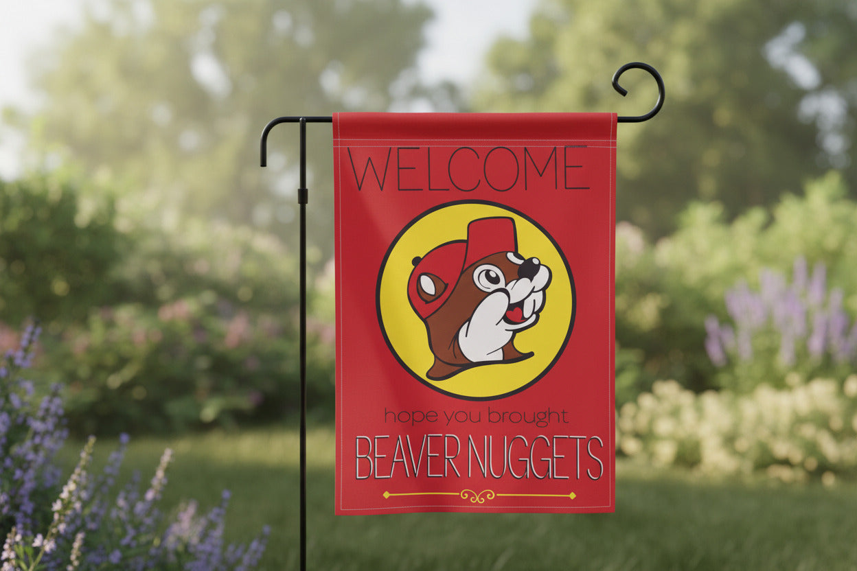 A picture of a pretty red garden flag standing up in a green garden containing lavender flowers and green, fully mature trees.  The front of the flag has a  Buc-ee's logo dead in the center - a cheerful beaver with an overbite, wearing a red cap, framed by a yellow circle.  The top of the flag reads WELCOME in thin uppercase black text.  The bottom of the flag reads "hope you brought" in lowercase black text, then "BEAVER NUGGETS" in white/black uppercase text.  