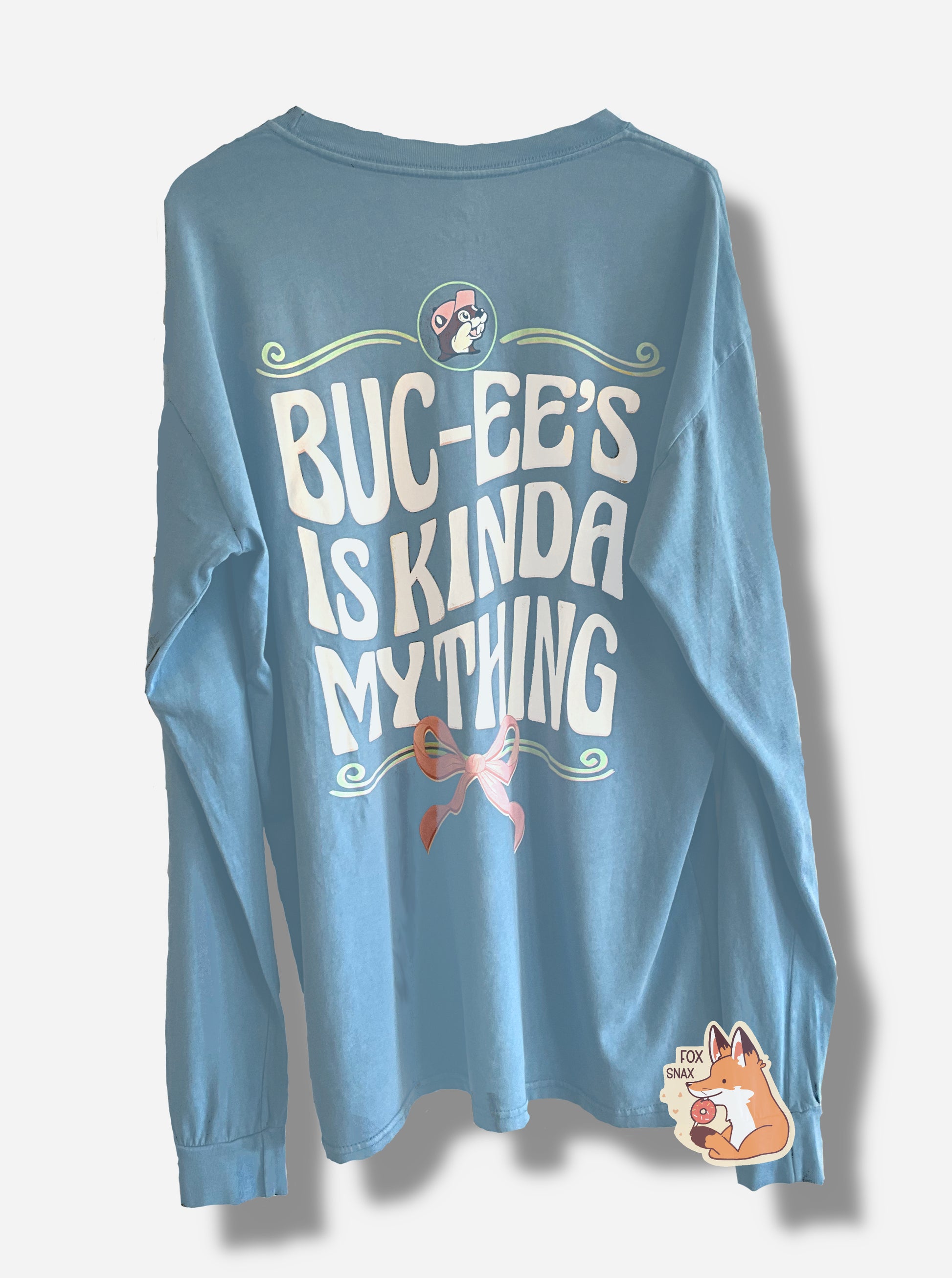 A picture of the back of a light blue, long sleeve shirt.  The top of the shirt has the Buc-ee's logo at the top, followed by the text "BUC-EE'S IS KINDA MY THING" in flowing white script.  The bottom has a tied pink bow at the bottom of it.