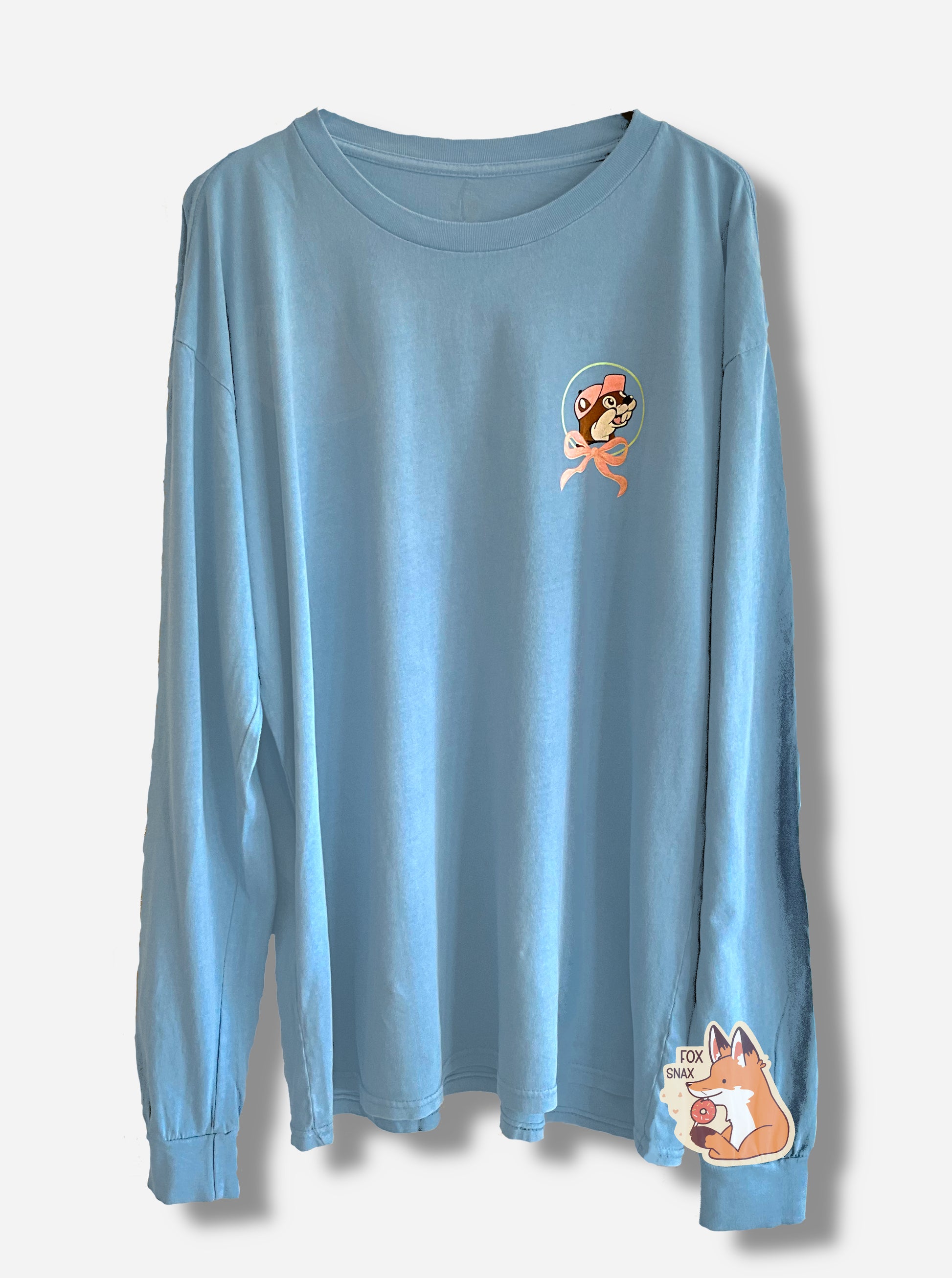 A picture of a light blue, long sleeve shirt.  The upper left of the shirt has the Buc-ee's logo, framed by a tied pink bow at the bottom of it, and Buc-ee's hat is in pink.