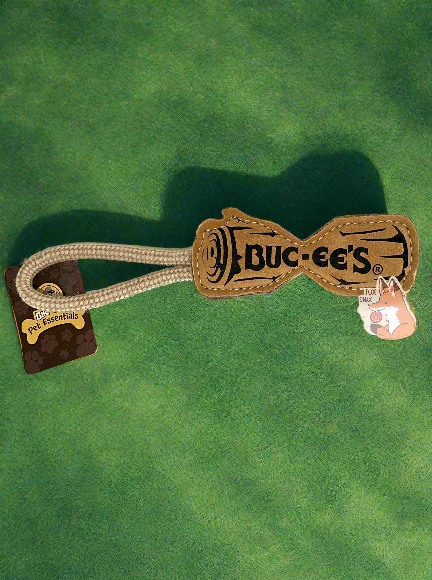 A picture of a Buc-ee's "Pet Essentials" Log Rope dog toy.  The body of the toy is shaped like a log - roughly - with a flat body and a dip in the middle, presumably to make it easier for a dog to bite it.  On one end is a strong tan rope.  The body of the tool is tan, and also has a black outline of a log with BUC-EE'S written in the middle.  Both ends of the  rope are tied into one end of the "log", making a loop for a person to hold on to. The background is of very short-cropped grass in a backyard.