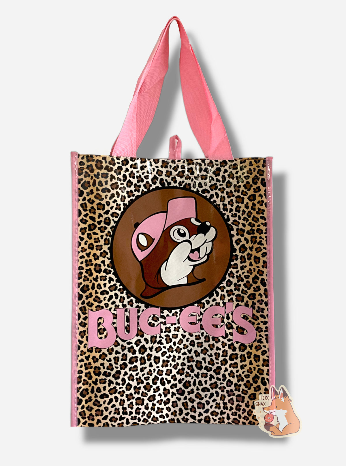 A picture of a large rectangular pink plastic shopping bag with the Buc-ee The Beaver logo on it.  Buc-ee has a light pink hat, and the background behind him is dark brown, and he's surrounded by a tan and brown leopard spots.  The bag itself reads BUC-EES in pink text.  The strap on the bag is made of pink cloth.  The vertical edges of the bag have a thin pink stripe.