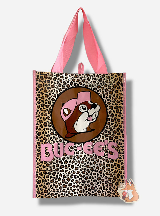 A picture of a large rectangular pink plastic shopping bag with the Buc-ee The Beaver logo on it.  Buc-ee has a light pink hat, and the background behind him is dark brown, and he's surrounded by a tan and brown leopard spots.  The bag itself reads BUC-EES in pink text.  The strap on the bag is made of pink cloth.  The vertical edges of the bag have a thin pink stripe.