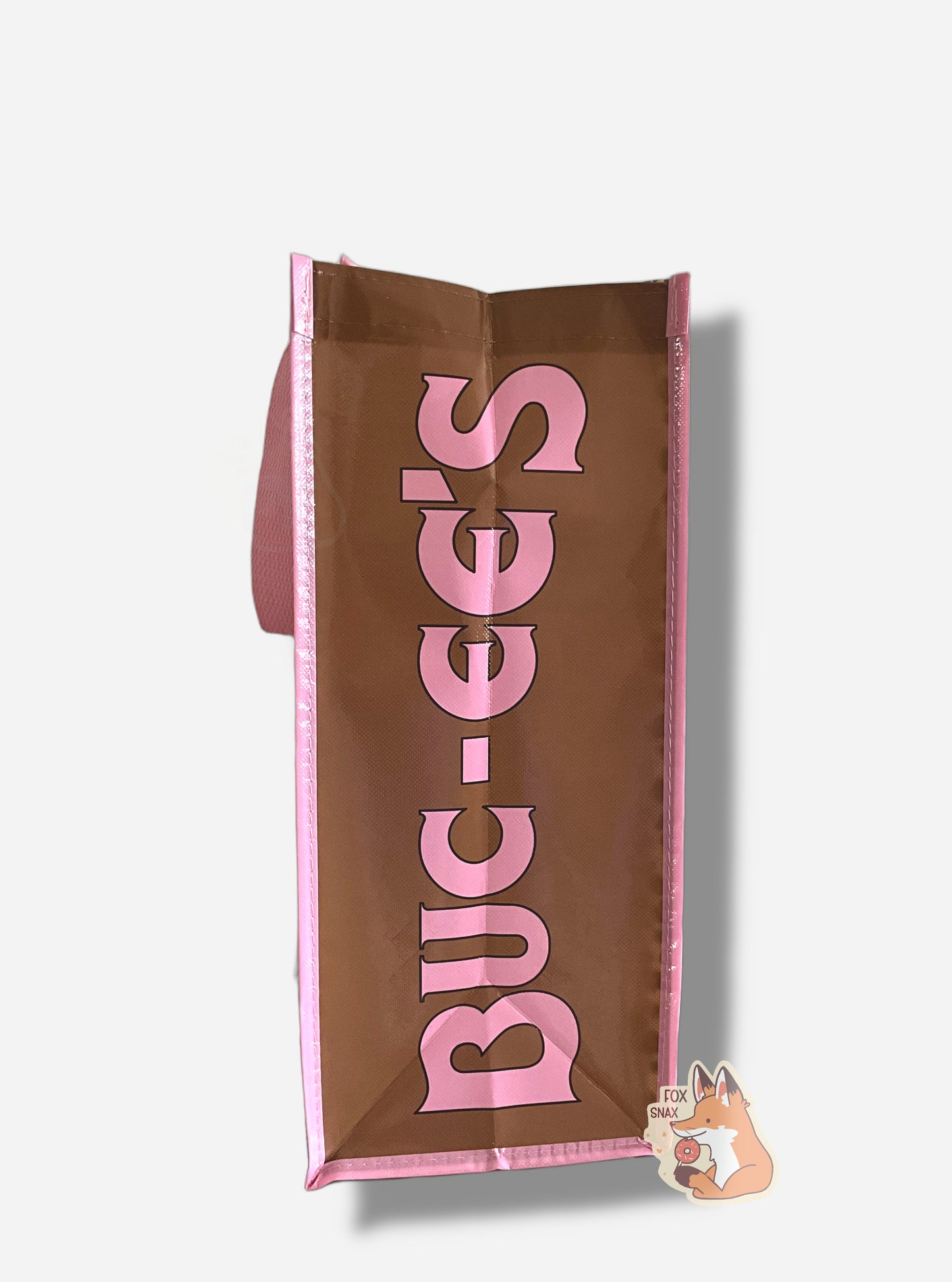 A picture of the side of a large rectangular pink and brown plastic shopping bag.  The bag itself reads BUC-EES in pink text on the side, and the background is brown.  The strap on the bag is made of pink cloth.  The vertical edges of the bag have a thin pink stripe.