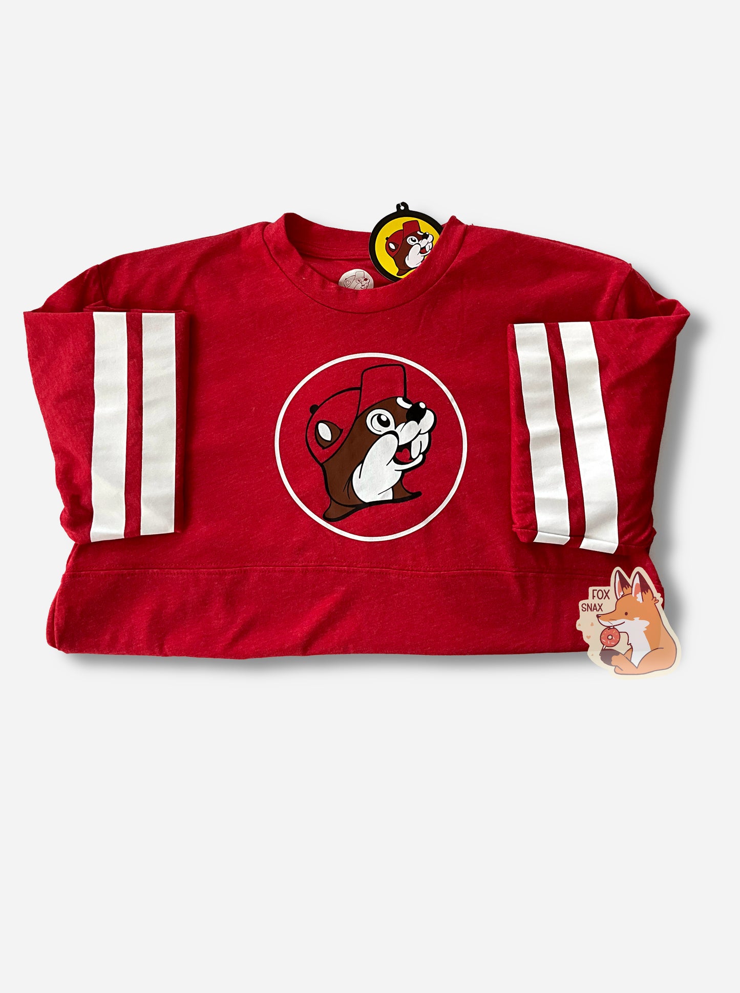 A picture of a long sleeve varsity shirt in red, with two thick white stripes around each of the upper arms.  The Buc-ee the beaver logo is in the middle, with a red ball cap, against a red circle with a white surround. In this picture, the arms are folded.  