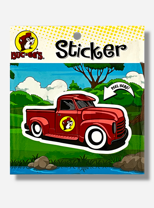 A picture of a Buc-ee's vinyl sticker.  The sticker is of a shiny but old model red pickup truck.  On the passenger side door of the truck is the Buc-ee's logo, which which is a cheerful beaver with a red ball cap.  The background of the sticker reads STICKER, and shows a countryside scene, complete with trees, bushes, and a nearby riverbank with blue water.