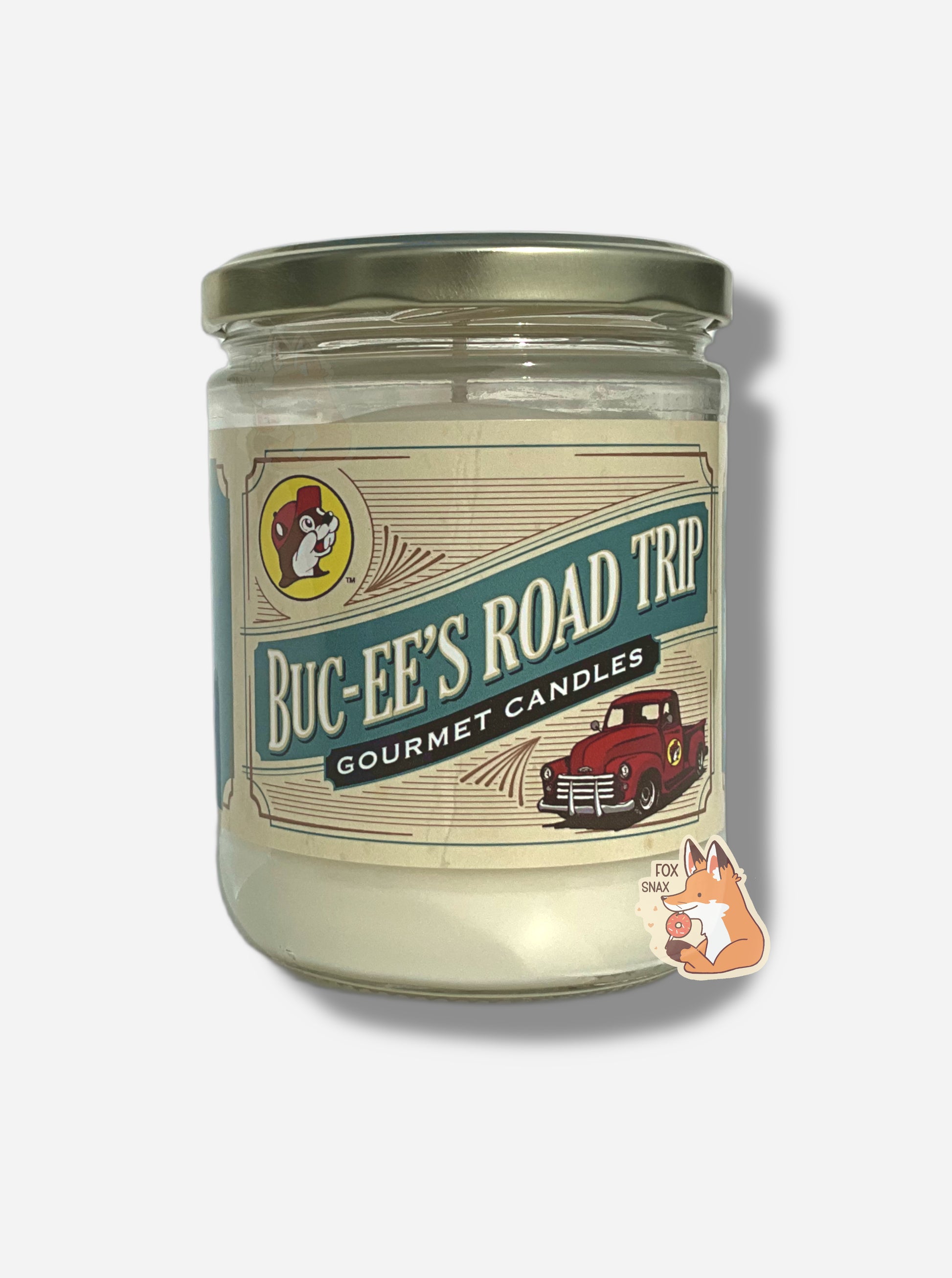 A picture of a white/yellow candle in a cylindrical glass container.  The lid of the container is a light gold colored metal lid.  The front of the candle has a yellow sticker with the Buc-ee's logo, then underneath that, BUC-EE'S ROAD TRIP, then GOURMET CANDLES, then a picture of a old-style red pickup truck.
