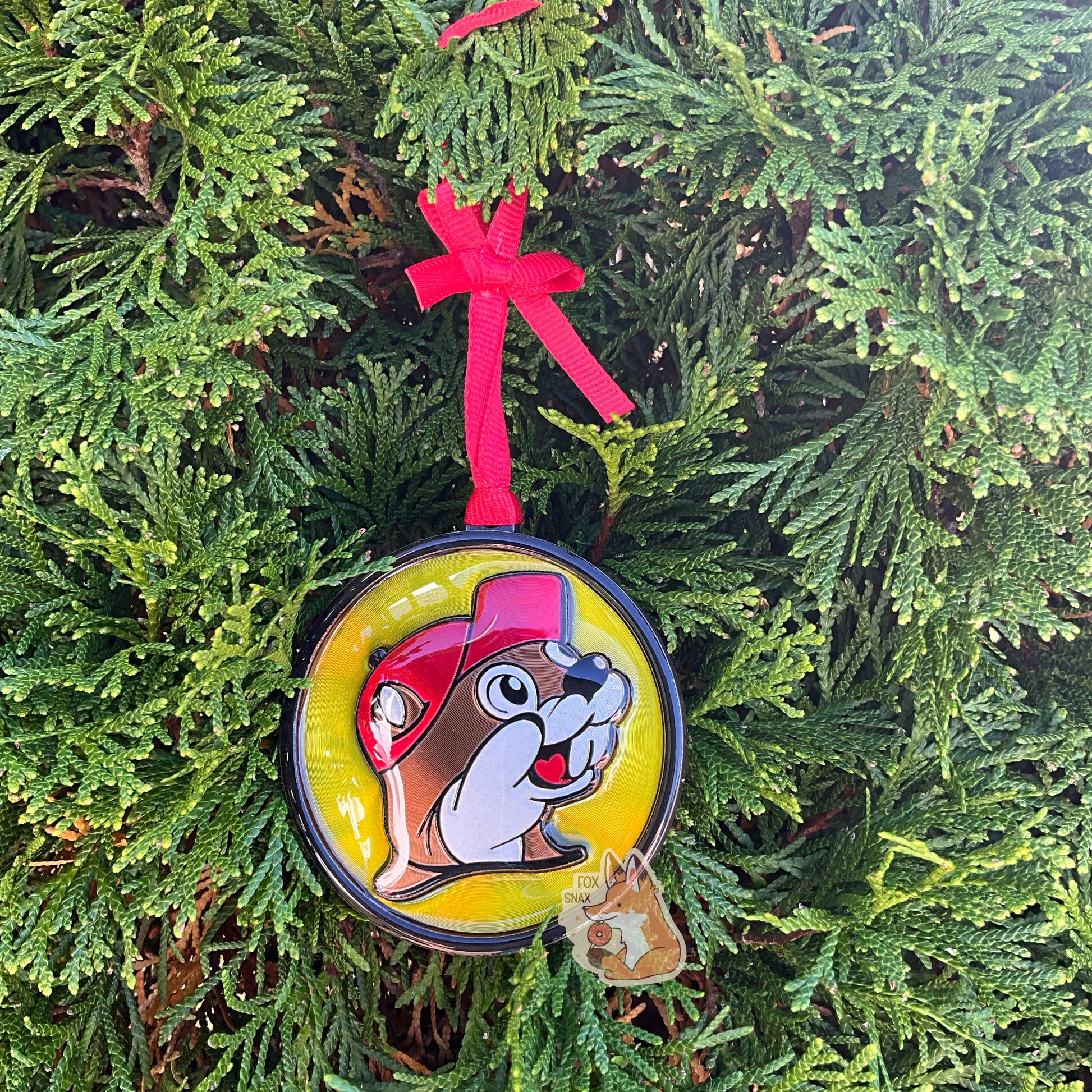 A picture of the front of a round, shiny, Christmas ornament.  The ornament is of the Buc-ee's logo - a smiling beaver wearing a red ballcap.  It is made out of foil and plastic, and is extremely shiny.  A red ribbon pokes out from the top of the ornament's body, tying it to the green evergreen branches of a pretty tree in the background.