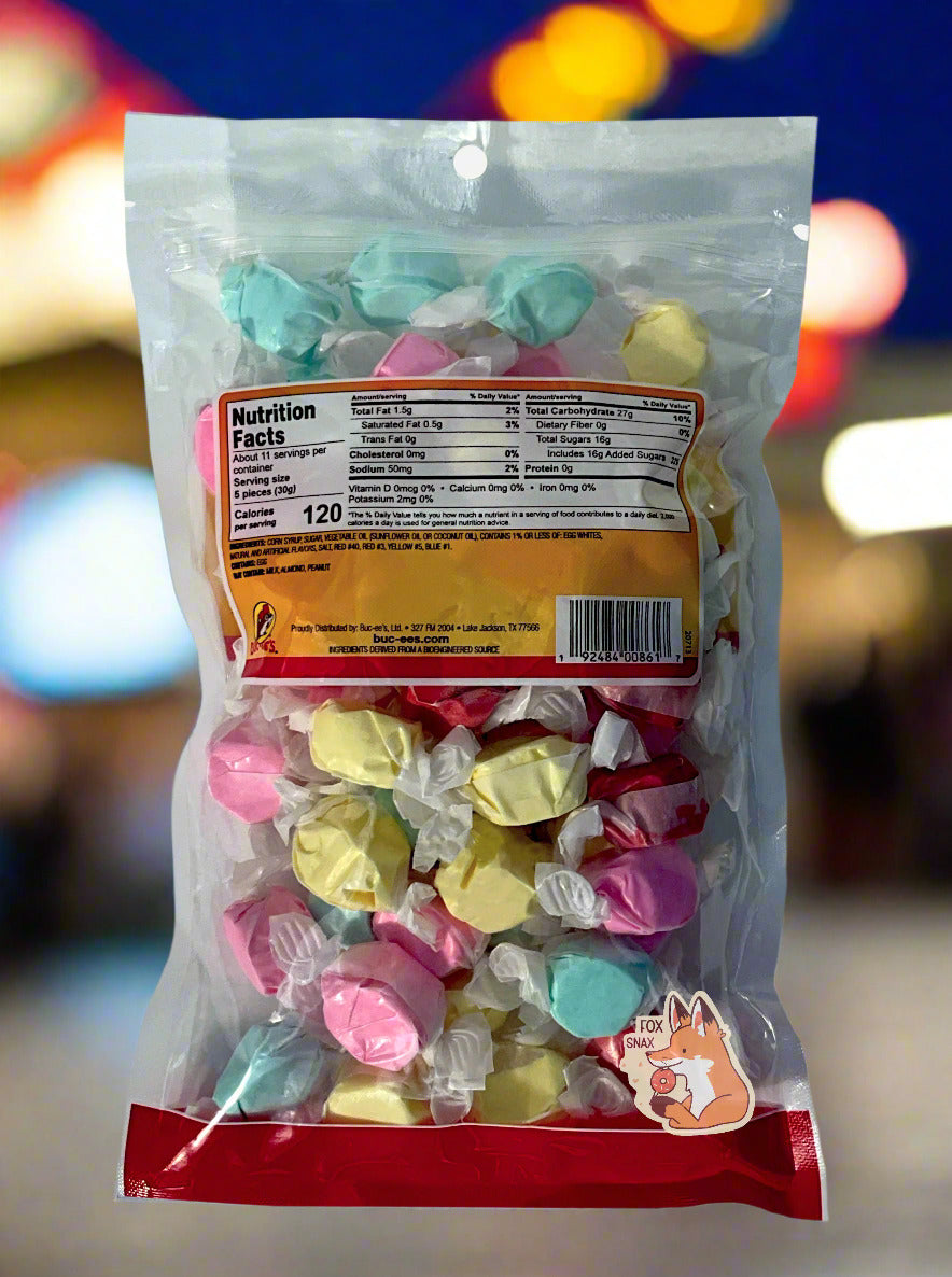 A picture of the back of a plastic bag of Buc-ee's State Fair Salt Water Taffy.  The bag is clear plastic.  Inside it, twists of candy in four different wrapping colors (cotton candy, buttered popcorn, bubble gum, red licorice) can be seen. Bright colorful fair lights at night can be seen behind the bag.