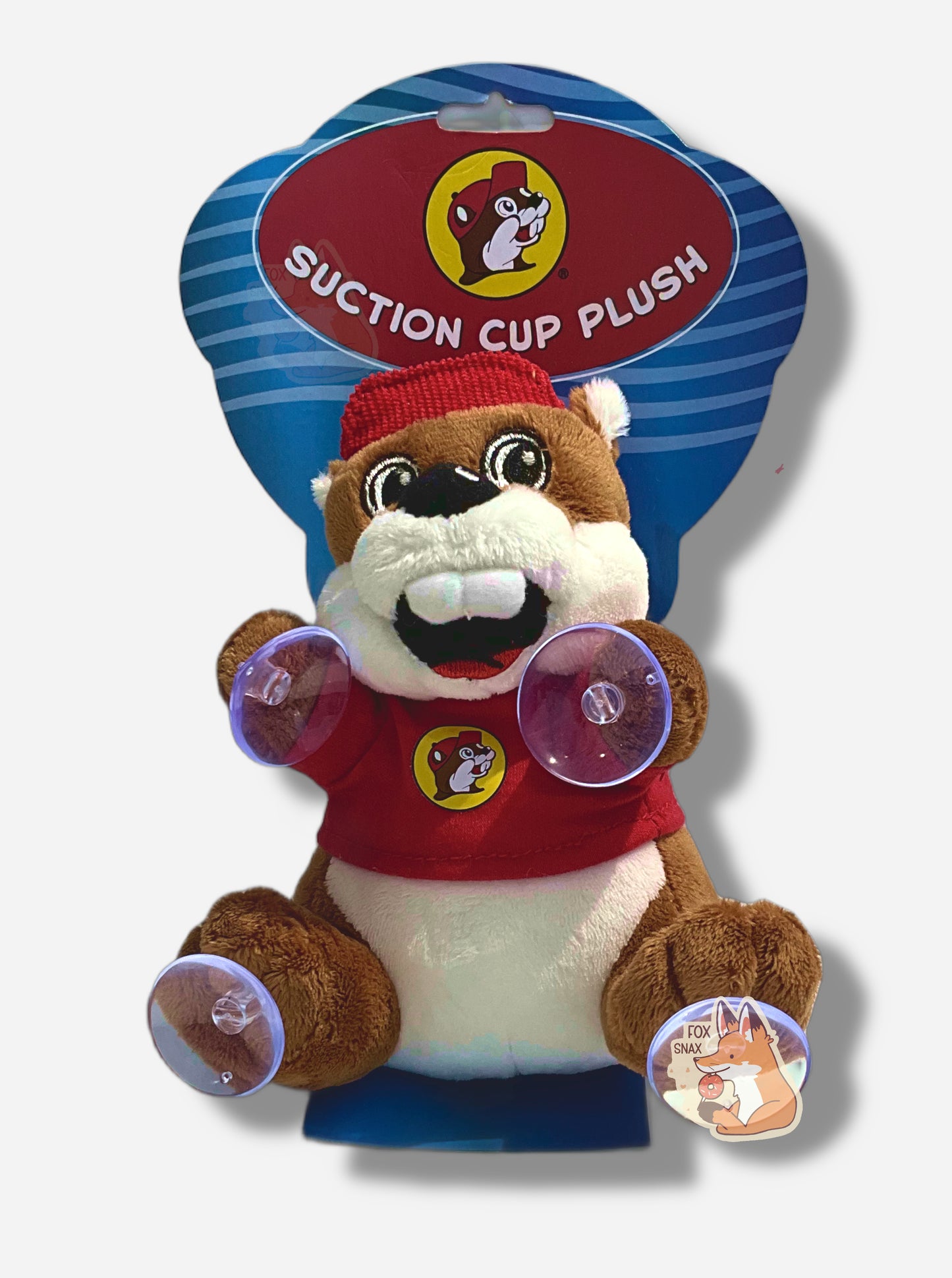 A picture of an adorable Buc-ee the Beaver plush.  The plush is of Buc-ee, wearing a red Buc-ee's shirt and red ball cap, and is looking to the left slightly.  On each paw, he has a small clear suction cup; he's intended to be placed on a car window.  He is roughly a six inches tall and covered in plush brown and white stuffed animal "fur".  Behind him is a paper wrapper in blue and red that reads "Suction Cup Plush"