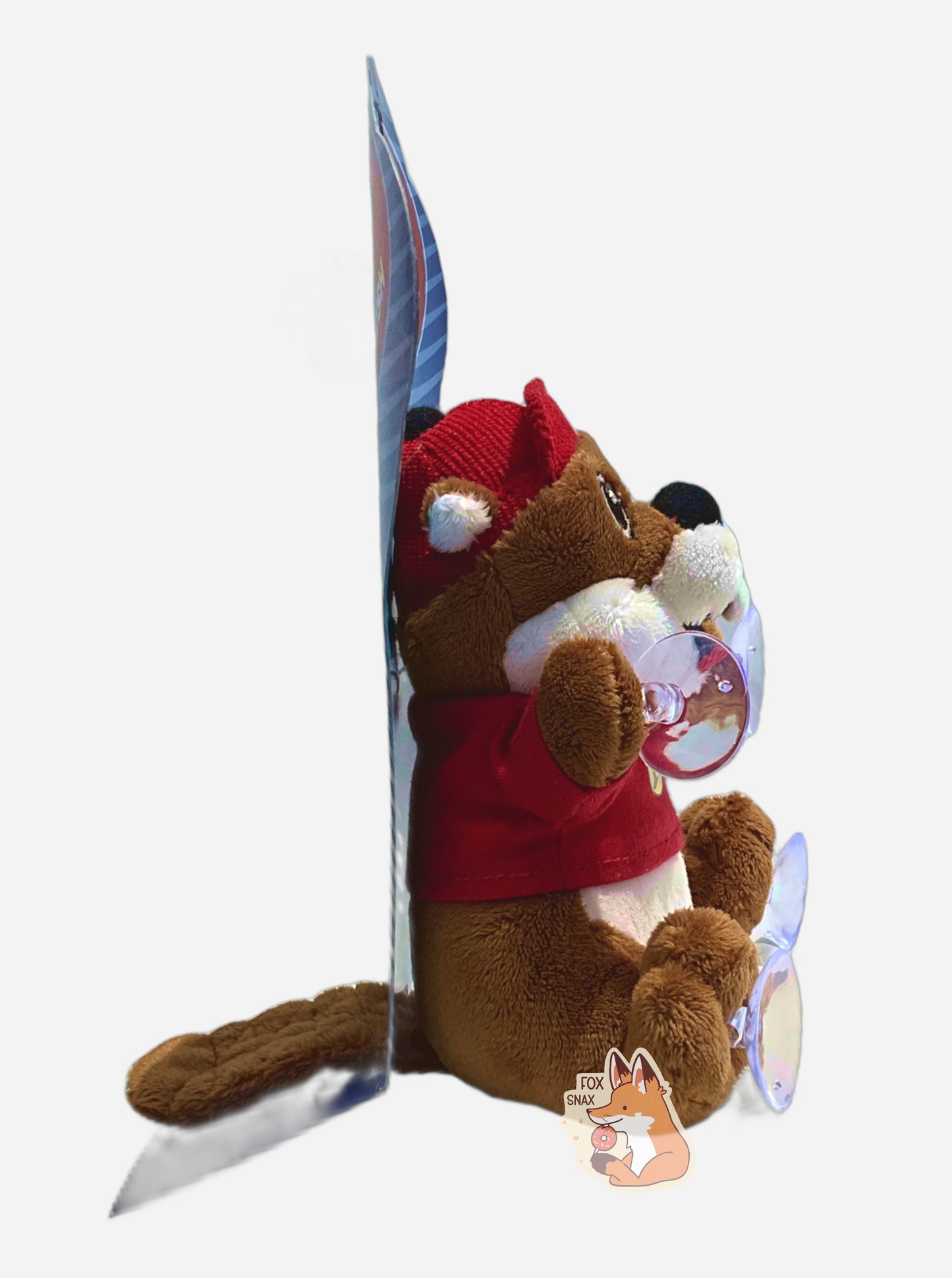 A side view of of an adorable Buc-ee the Beaver plush.  The plush is of Buc-ee, wearing a red Buc-ee's shirt and red ball cap.  On each paw, he has a small clear suction cup; he's intended to be placed on a car window.  He is roughly six inches tall and covered in plush brown and white stuffed animal "fur", with a brown beaver tail poking from his backside.