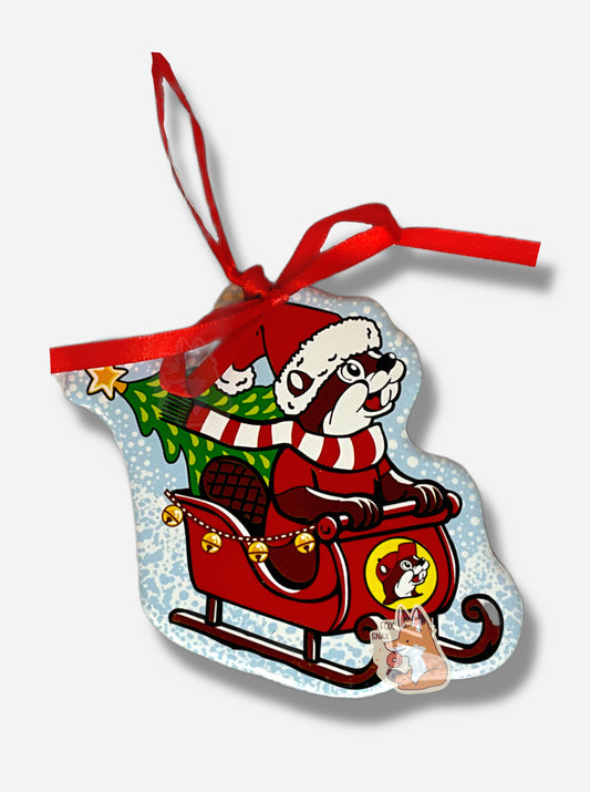 A picture of the front of a ceramic ornament, with a red ribbon at the top to hang it on a tree.  The ornament has a picture of Buc-ee The Beaver on it, but Buc-ee is wearing a Santa hat instead of his usual red ball cap, a red and white striped scarf, and a red shirt.  He is holding on to the front of a red sleigh with brown runners.  In the back of the sleigh is a green Christmas tree topped with a yellow star.  Jingle bells decorate the side of the sleigh, and the background is a light blue/white.