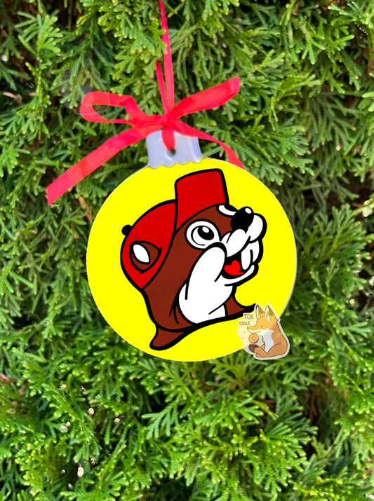 A picture of the front of a ceramic, mostly round ornament, with a red ribbon at the top to hang it on a tree.  The ornament has a picture of Buc-ee The Beaver on it, the same as the gas station logo one could see from the road.  The background of the ornament is solid yellow.  This ornament is hanging from an evergreen tree.