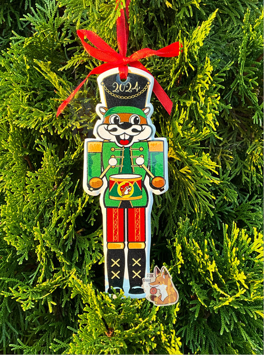 A picture of Buc-ee the Beaver, dressed as a nutcracker.  Buc-ee's hat is a large black one and has 2024 on it.  In each hand, he is holding a drumstick, and a small drum (with a Buc-ee's logo) in green in red is at his waist.  His legs are covered in ornate red pants with gold trim; his boots are large and black.  His vest is green with yellow buttons.  His smile is framed with a black mustache.  A red ribbon ties the ornament to a Christmas tree in the background of the picture.