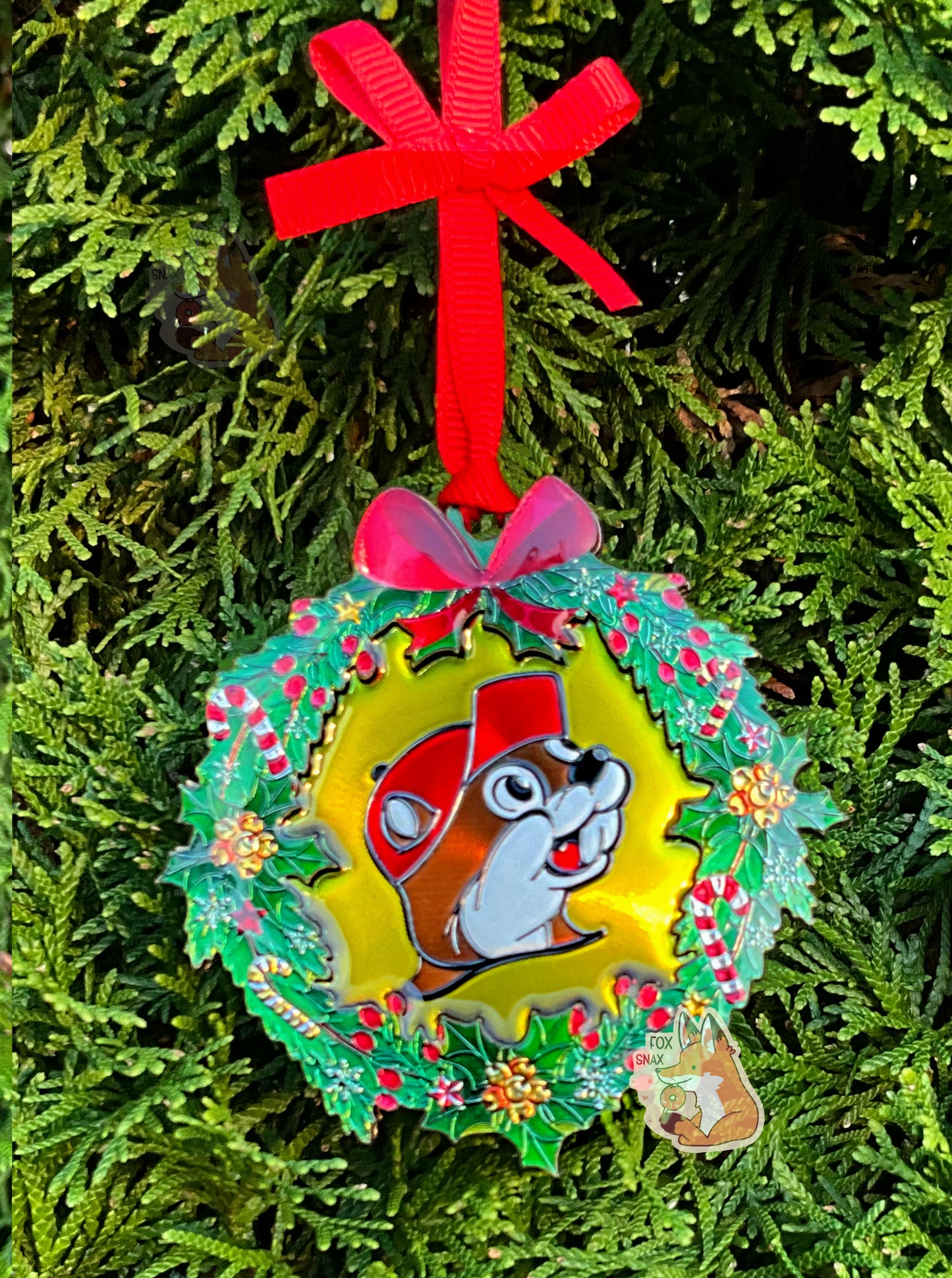 A picture of a circular ornament, in the shape of a green wreath.  The wreath is decorated with candy canes, holly berries, and yellow bells.  In the center of the ornament is the Buc-ee's logo (also in foil).  The top of the ornament has a foil red bow on it, and is held up by a bright red bow to an evergreen tree in the background.