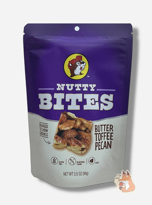 A picture of a white and purple bag of Buc-ee's Butter Toffee Pecans. The Buc-ee's logo can be seen in the top center of the bag, then under that, NUTTY BITES.  In the center of the bag is a brown square of Nutty Bite, covered in butter toffee and pecans. To the left of the bite reads text, "ENLARGED TO SHOW GOODNESS".  To the right of the bite reads text, "BUTTER TOFFEE PECAN".  On the bottom of the bag it says GLUTEN FREE, then NO ARTIFICIAL FLAVORS, then VEGAN.  3.5 oz.