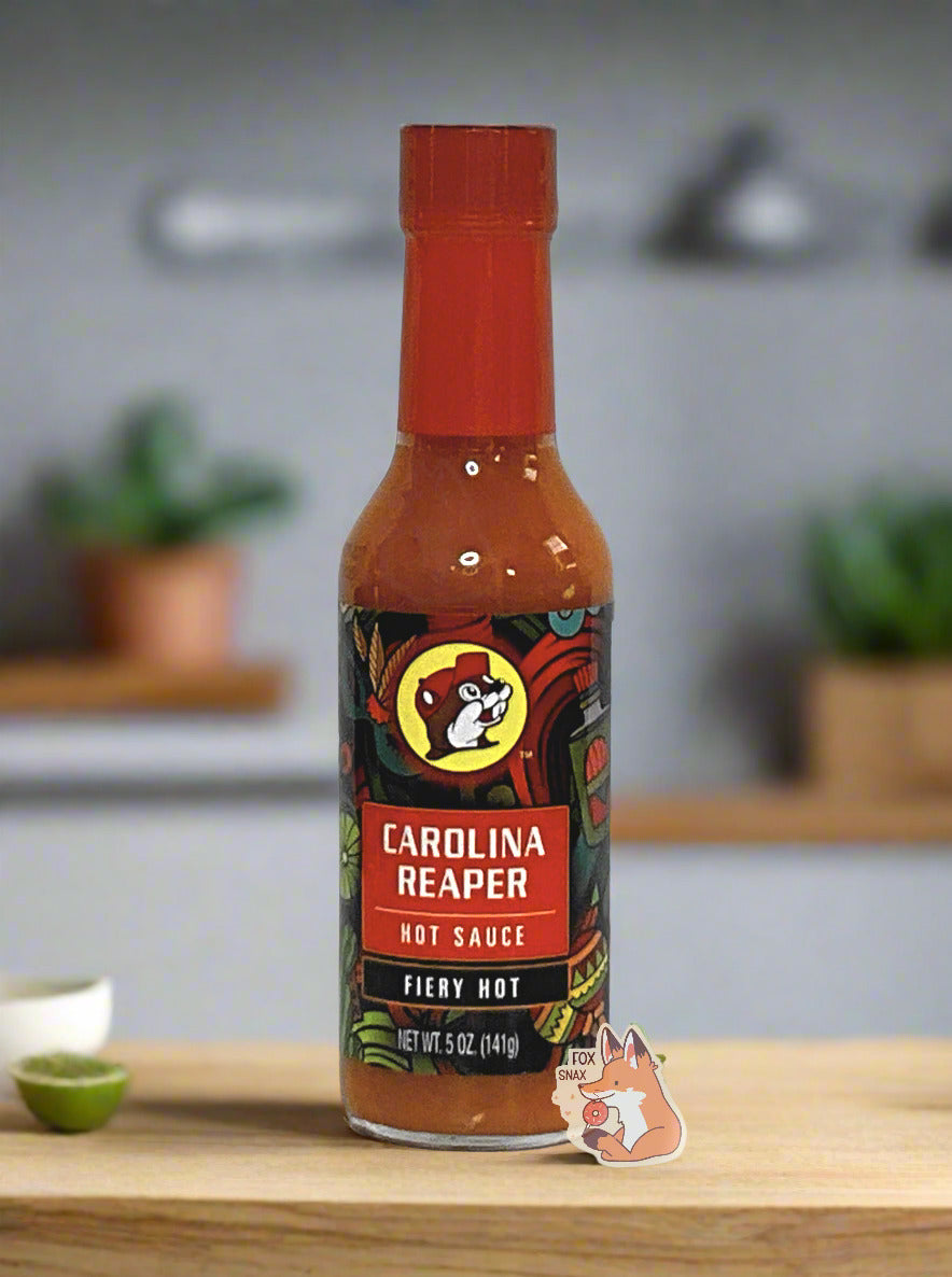 A picture of a small, tall glass bottle of hot sauce.  The top of the bottle is sealed with a red plastic cover on a black cap. The front label of the bottle reads CAROLINA REAPER HOT SAUCE, FIERY HOT.  The Buc-ee's logo can be seen on the front; the background is a fairly busy abstract artwork featuring a cactus, two maracas, a lime, and a Mardi Gras mask.  The sauce inside is red/orange.  The bottle is sitting on a countertop, a cut lime is nearby, as is a white bowl.  The background is of a kitchen.