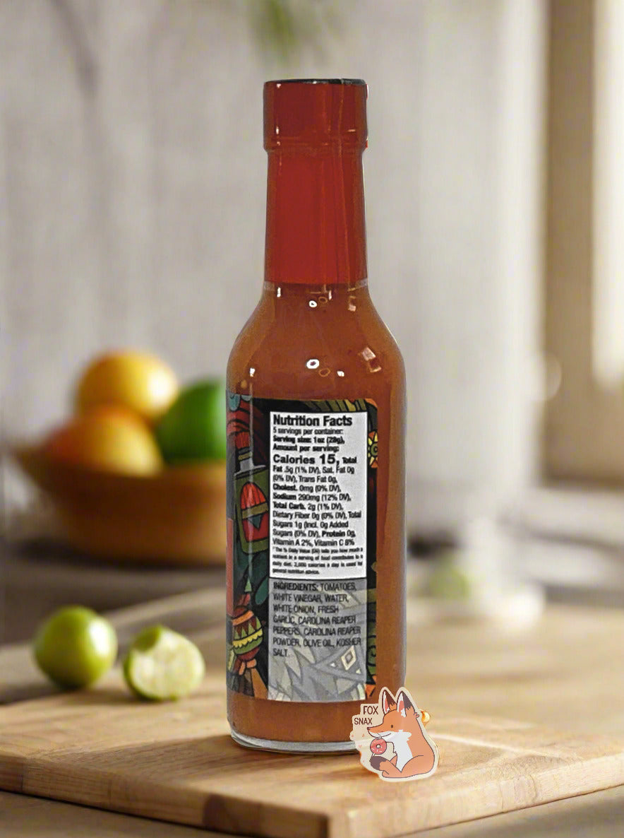 A picture of a small, tall glass bottle of hot sauce. The top of the bottle is sealed with a plastic top. The sauce inside is red/orange.  The bottle sits on a wooden cutting board; cut limes can be seen in the kitchen background, as can oranges.
