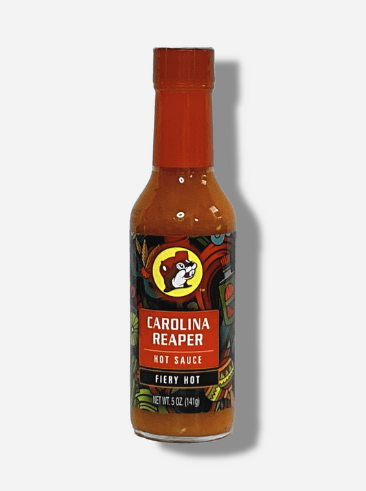 A picture of a small, tall glass bottle of hot sauce.  The top of the bottle is sealed with a shiny orange plastic top.  The front label of the bottle reads CAROLINA REAPER HOT SAUCE, FIERY HOT.  The Buc-ee's logo can be seen on the front; the background is a fairly busy abstract artwork featuring a cactus, two maracas, a lime, and a Mardi Gras mask.  The sauce inside is bright orange.