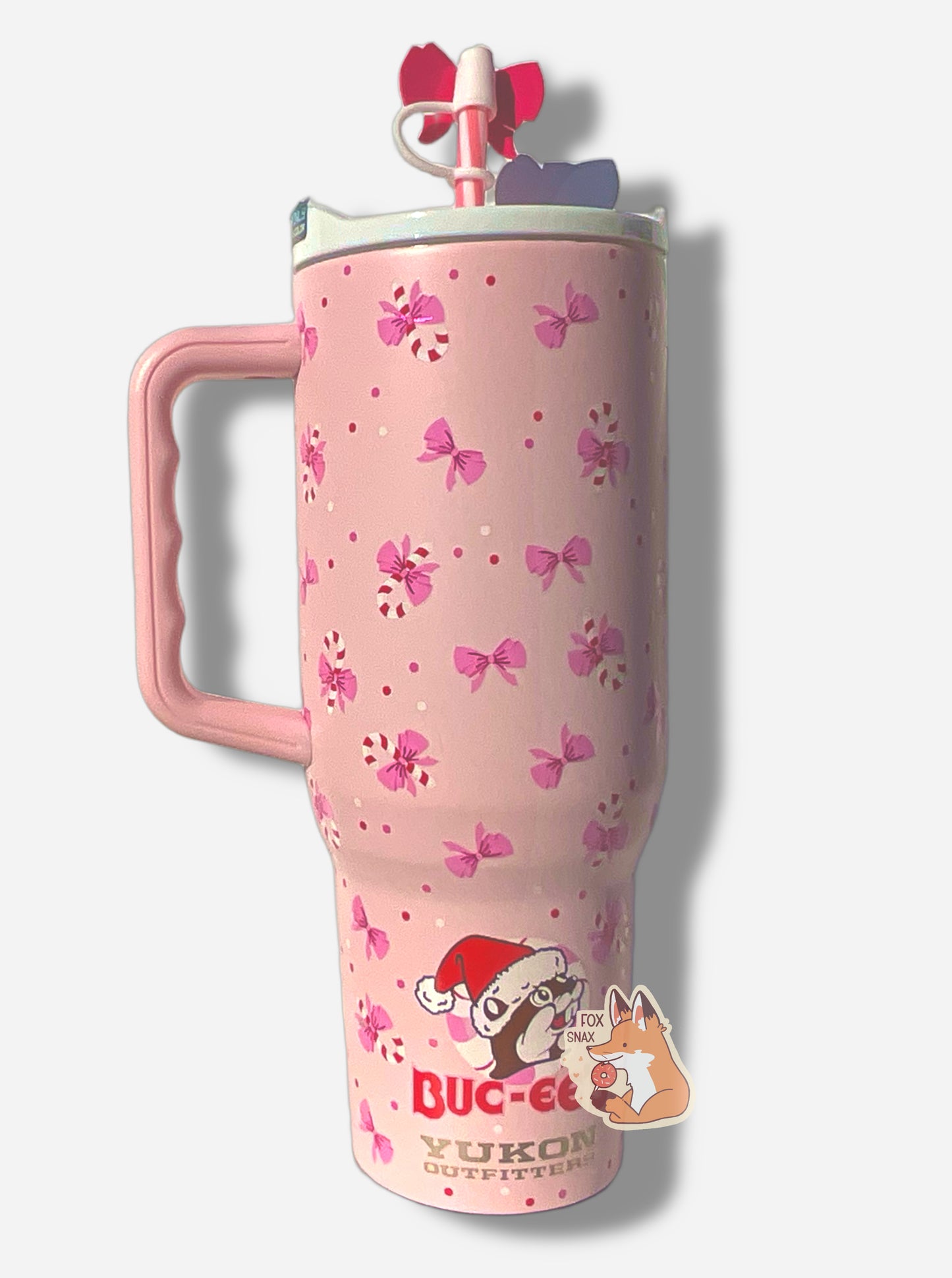A picture of a tall pink, skinny tumbler, with the Buc-ee's logoon the bottomBuc-ee is wearing a Santa cap, and is surrounded by a circular peppermint. The tumbler is paint covering stainless steel.  The handle, top, and pink peppermint colored straw are white and pink plastic. The lip of the tumbler is white;  background on the tumbler is pink, dotted with pink bows and candy canes!  The straw also has a pink bow on it.