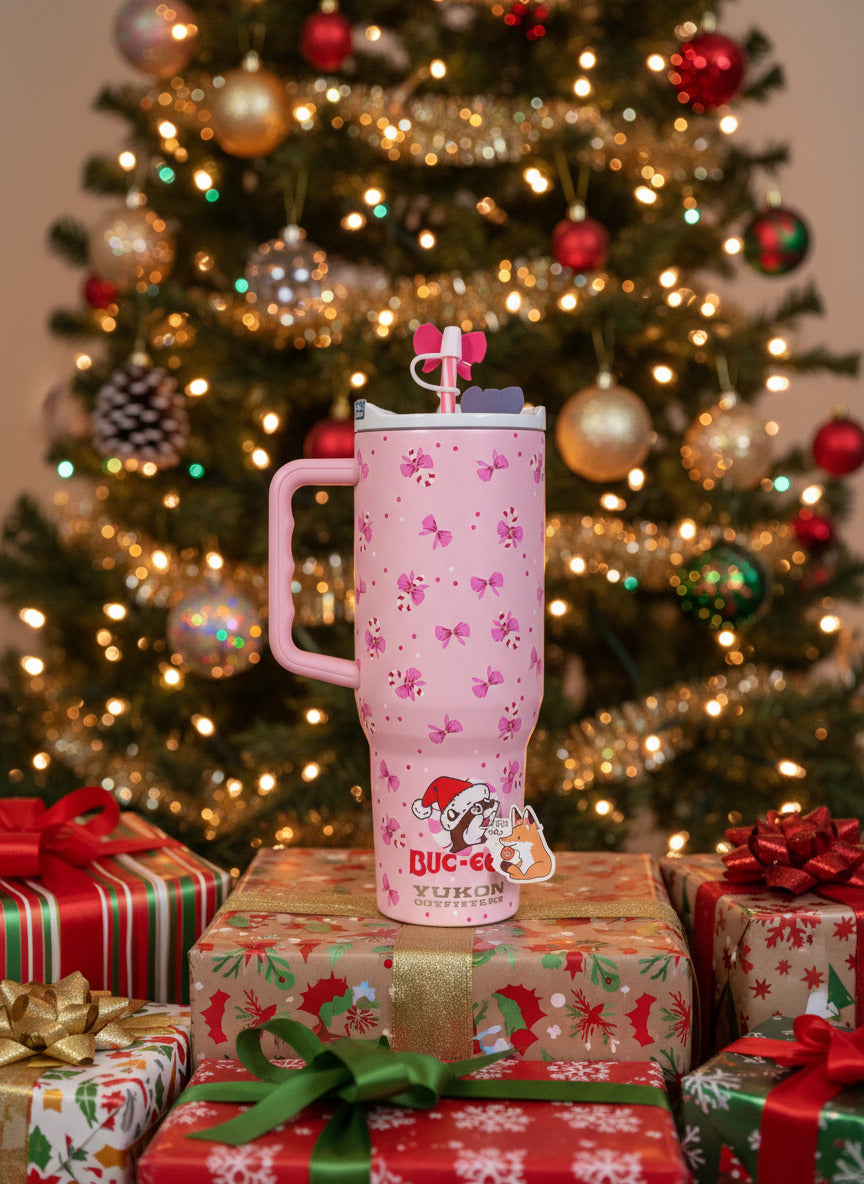 A picture of a tall pink, skinny tumbler, with the Buc-ee's logoon the bottomBuc-ee is wearing a Santa cap, and is surrounded by a circular peppermint. The tumbler is paint covering stainless steel.  The handle, top, and pink peppermint colored straw are white and pink plastic. The lip of the tumbler is white;  background on the tumbler is pink, dotted with pink bows and candy canes!  The straw also has a pink bow on it.  The tumbler is sitting on a pile of presents under a decorated Christmas tree.