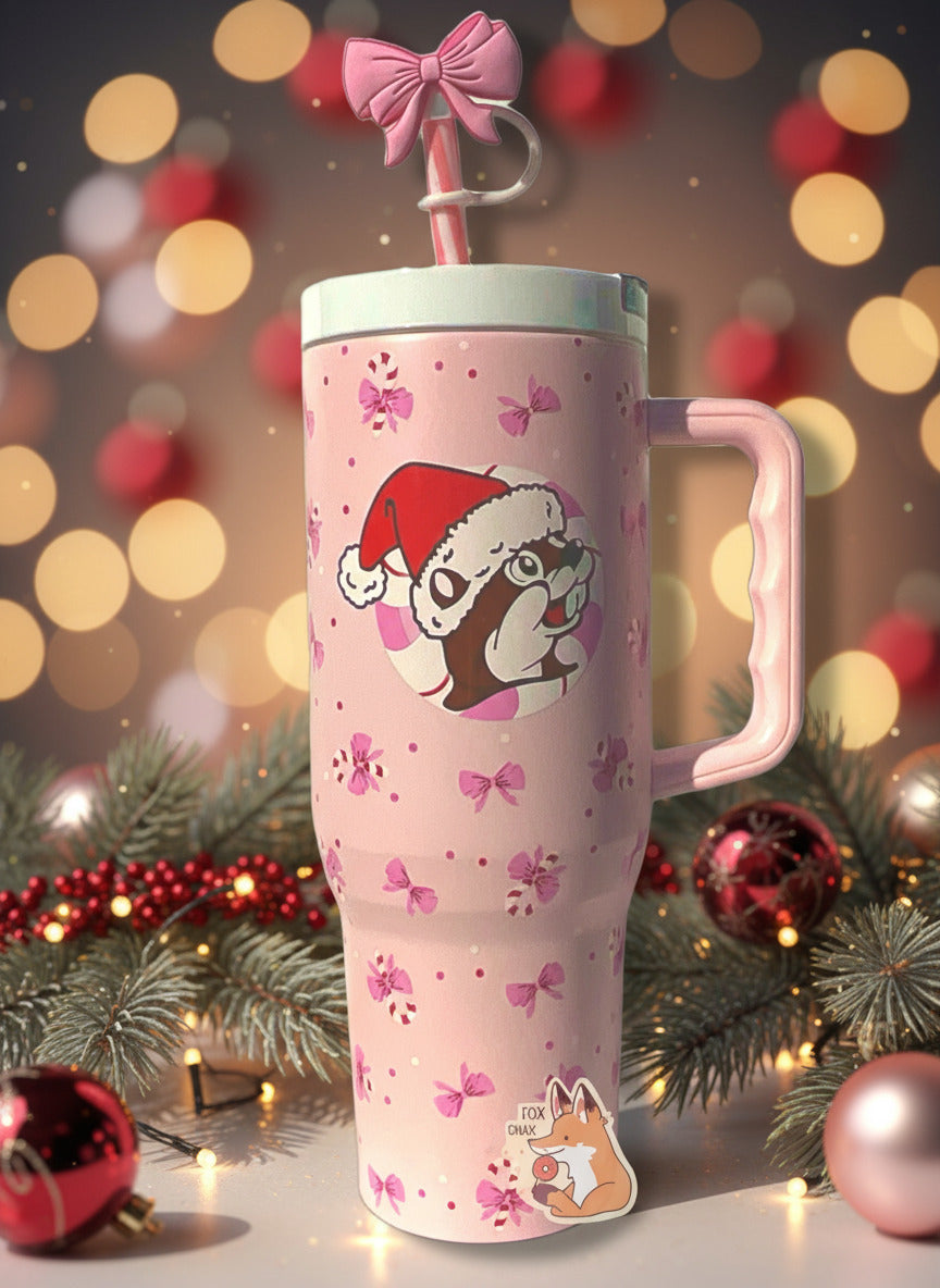 A picture of a tall pink, skinny tumbler, with the Buc-ee's logo in the middle; Buc-ee is wearing a Santa cap, and is surrounded by a circular peppermint. The tumbler is paint covering stainless steel.  The handle, top, and pink peppermint colored straw are white and pink plastic. The lip of the tumbler is white;  background on the tumbler is pink, dotted with pink bows and candy canes!  The straw also has a pink bow on it.  The background is a table with Christmas garland and pretty ornaments and lights.