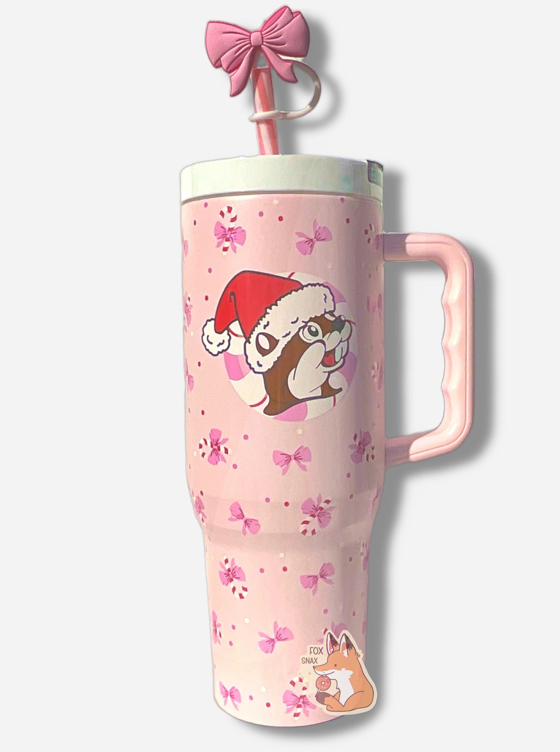 A picture of a tall pink, skinny tumbler, with the Buc-ee's logo in the middle; Buc-ee is wearing a Santa cap, and is surrounded by a circular peppermint. The tumbler is paint covering stainless steel.  The handle, top, and pink peppermint colored straw are white and pink plastic. The lip of the tumbler is white;  background on the tumbler is pink, dotted with pink bows and candy canes!  The straw also has a pink bow on it.