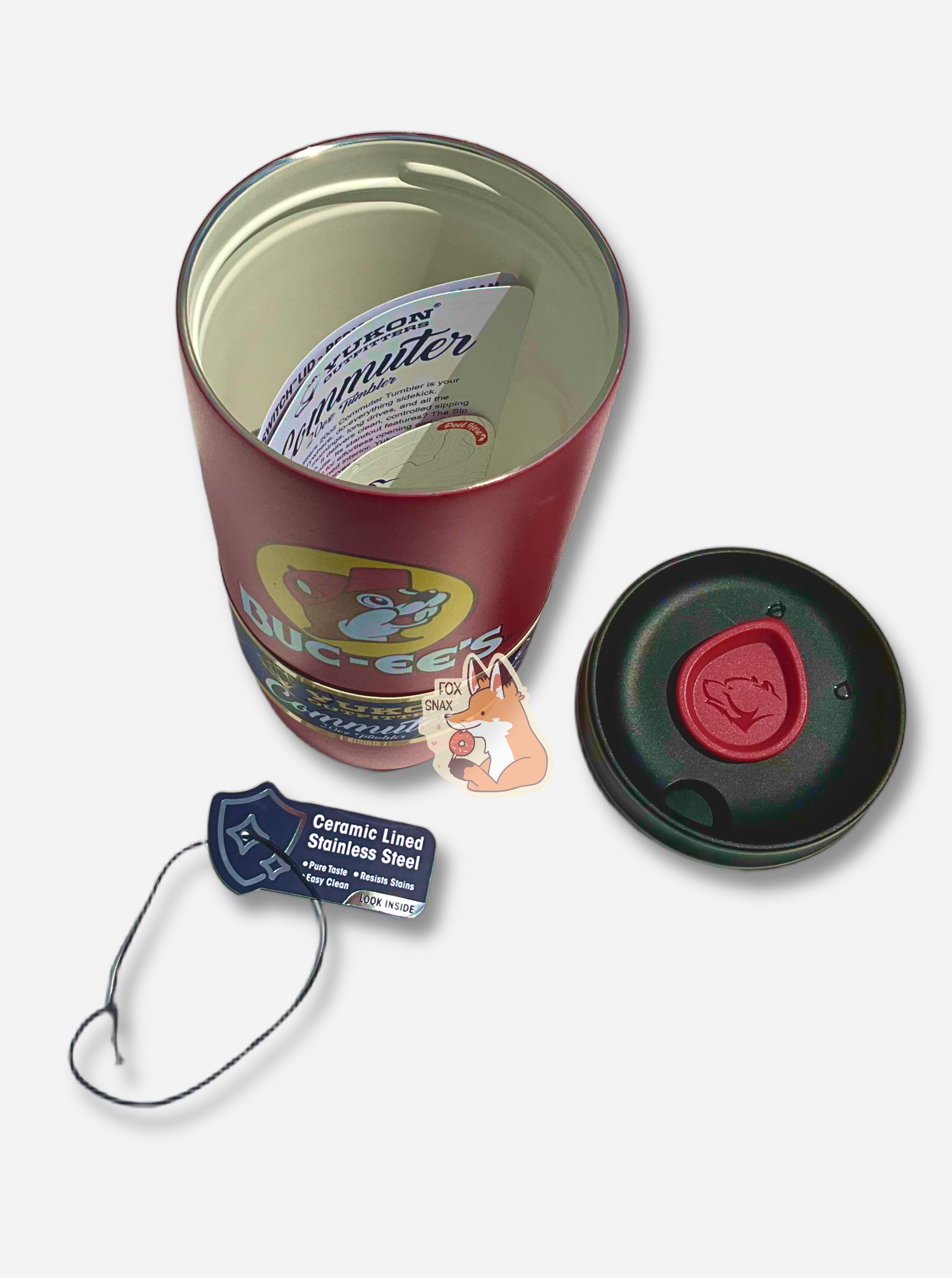 A three-quarters perspective picture of the top of a medium sized 20oz tumbler.  The surface of the cup is red paint over metal.  The top of the cup features a clear plastiic lid.  The top of the lid is removed; it's black plastic with a small sipping area on the exterior, near a red teardrop-shaped slider with the Yukon Outfitters logo (a grumpy bear) on top.  Inside the up, 'Commuter' instructions can be seen.  The interior of the cup features white ceramic lined stainless steel.