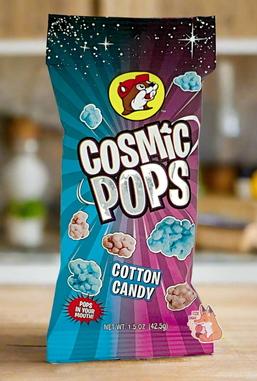A picture of Buc-ee's Cosmic Pops, in Cotton Candy flavor.  The bag starts with stars against a black background at the top, then shows rays of alternating pink and blue colors poking out of the center, to give the impression that the pops - lumpy odd shaped candies - are emerging from the center.  A logo on the bottom reads POPS IN YOUR MOUTH!  The background is of a kitchen; the bag is resting on a wooden kitchen table.