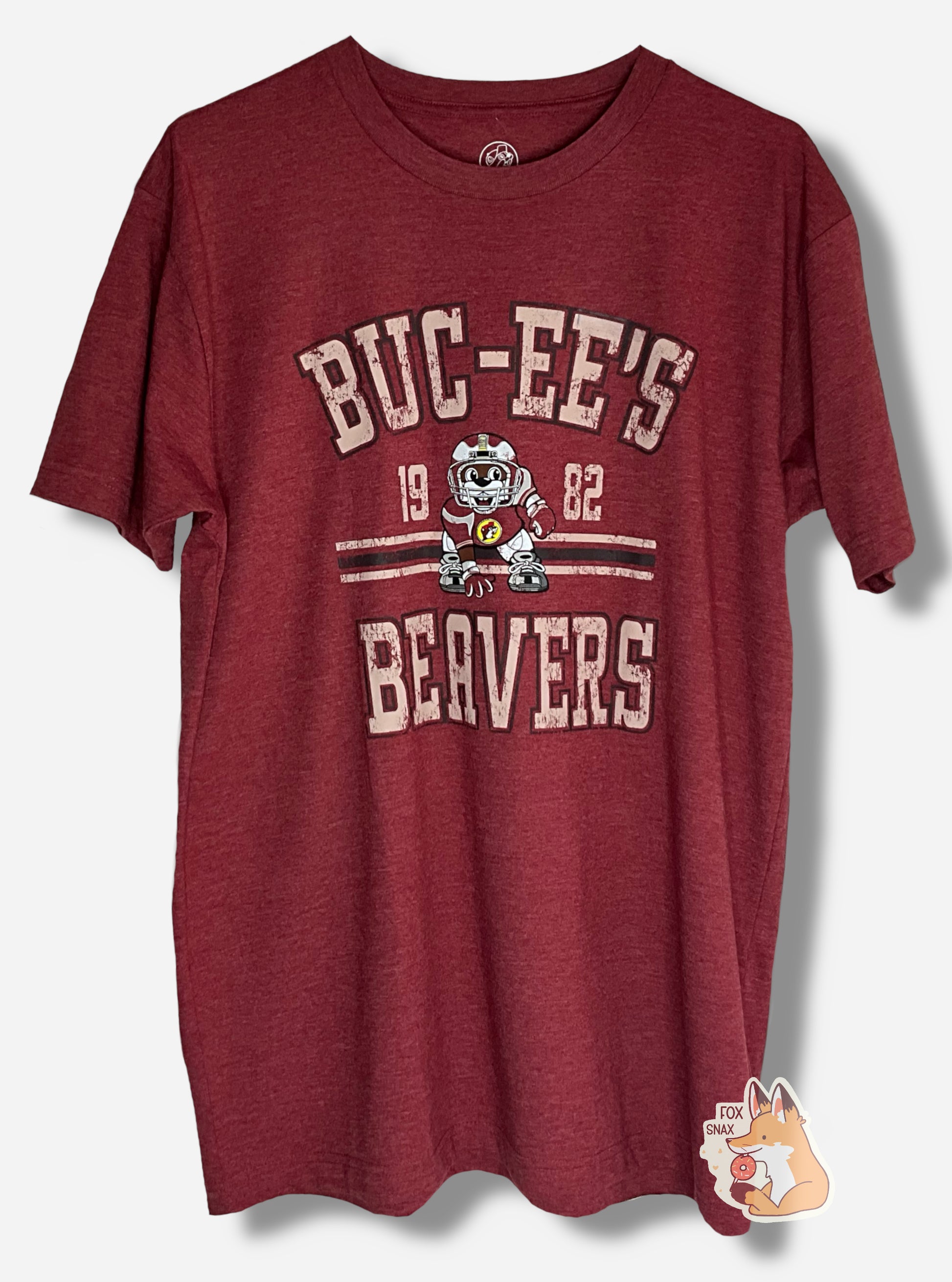 A picture of the front of a red t-shirt.  The shirt has text on the top that reads BUC-EE'S, and text on the bottom reading BEAVERS.  In the middle of the words are a picture of Buc-ee the Beaver wearing a dark red football uniform, complete with helmet.  On either side of Buc-ee are the numbers 19 and 82.